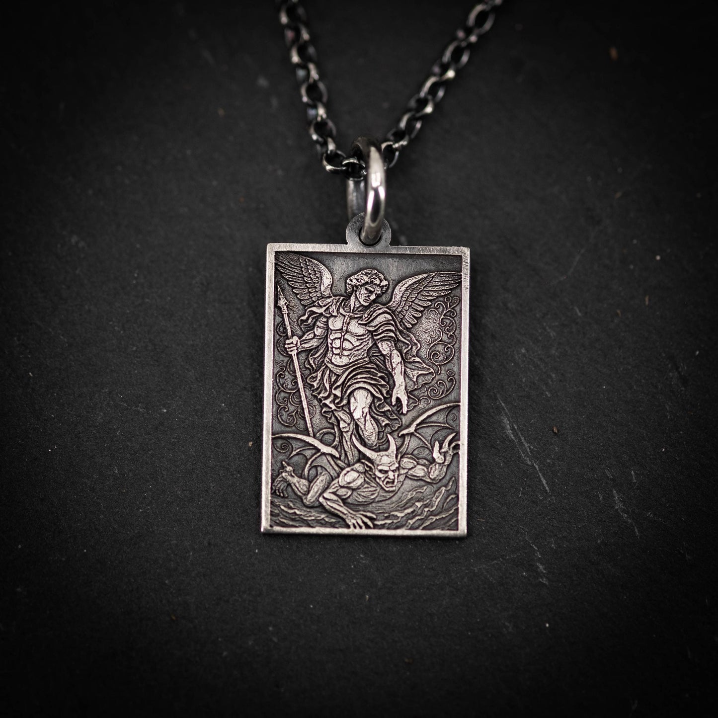 A rectangular pendant featuring an engraved image of a winged figure, possibly a saint or angel, with a battle scene in the background.