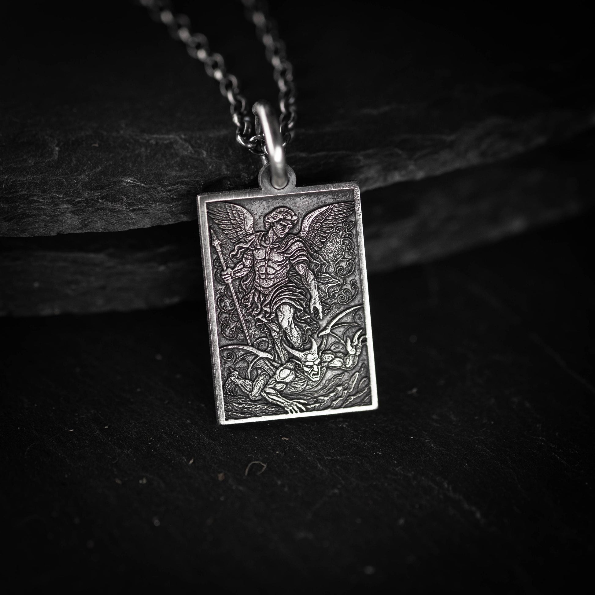 A rectangular pendant featuring an intricate engraved design, depicting a winged figure with a sword and shield, set against a dark background.