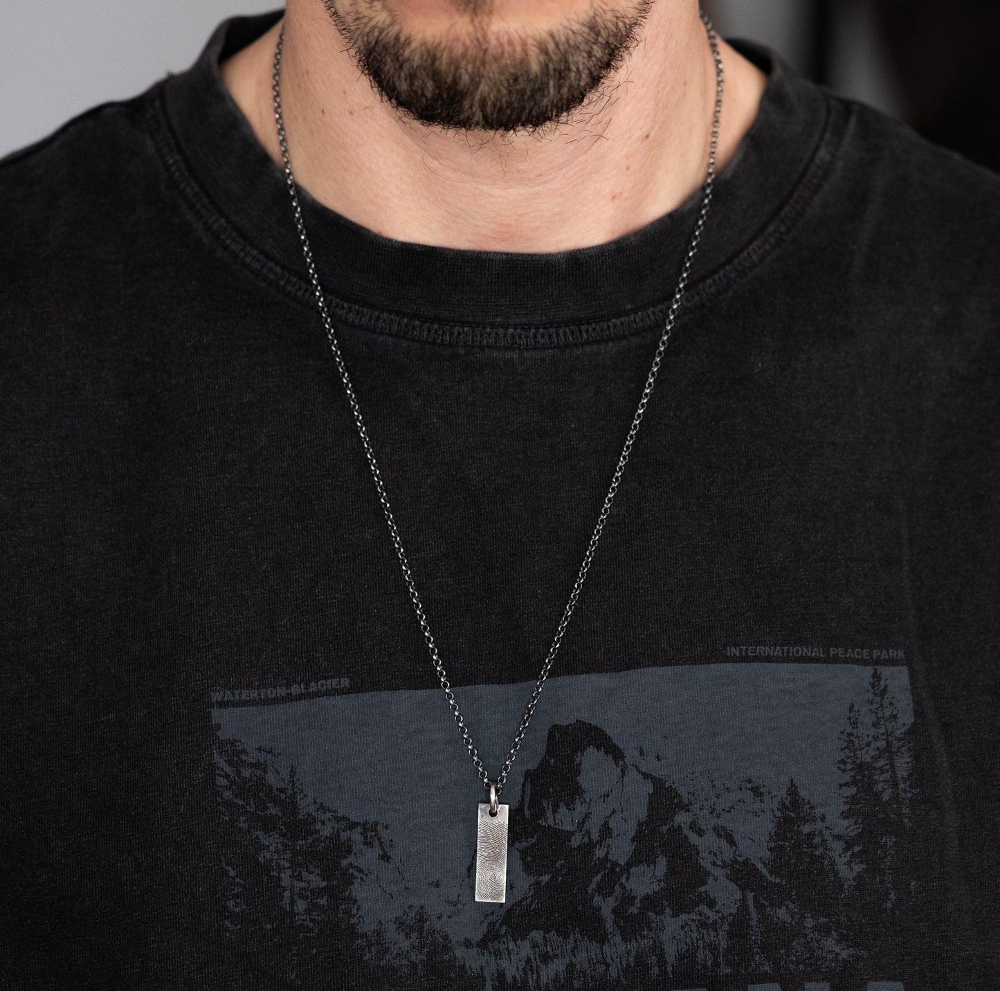 A close-up of a man wearing a black t-shirt with a graphic design and a silver necklace with a rectangular pendant.