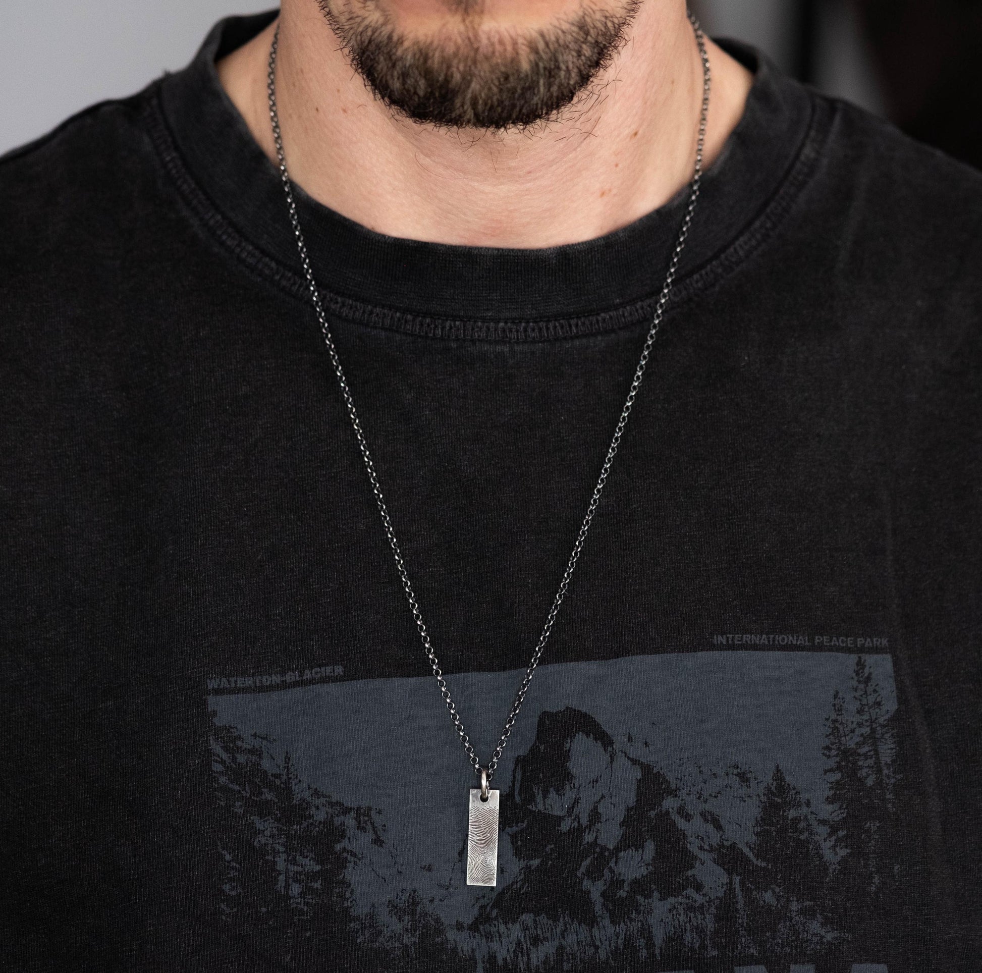 A close-up of a man wearing a black t-shirt with a graphic design and a silver necklace with a rectangular pendant.