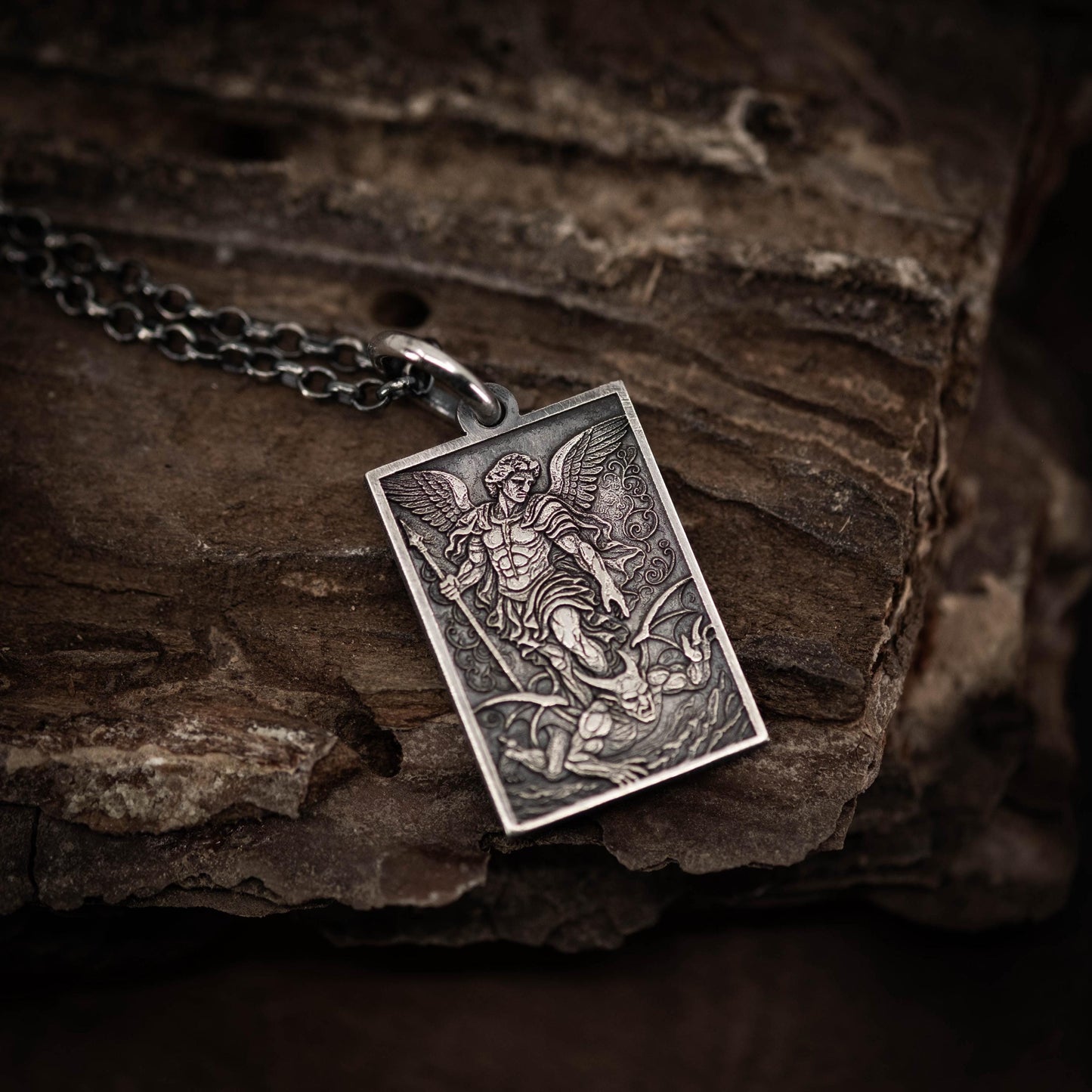 A silver pendant with an intricate design depicting a winged figure, possibly a saint or angel, is resting on a wooden surface.