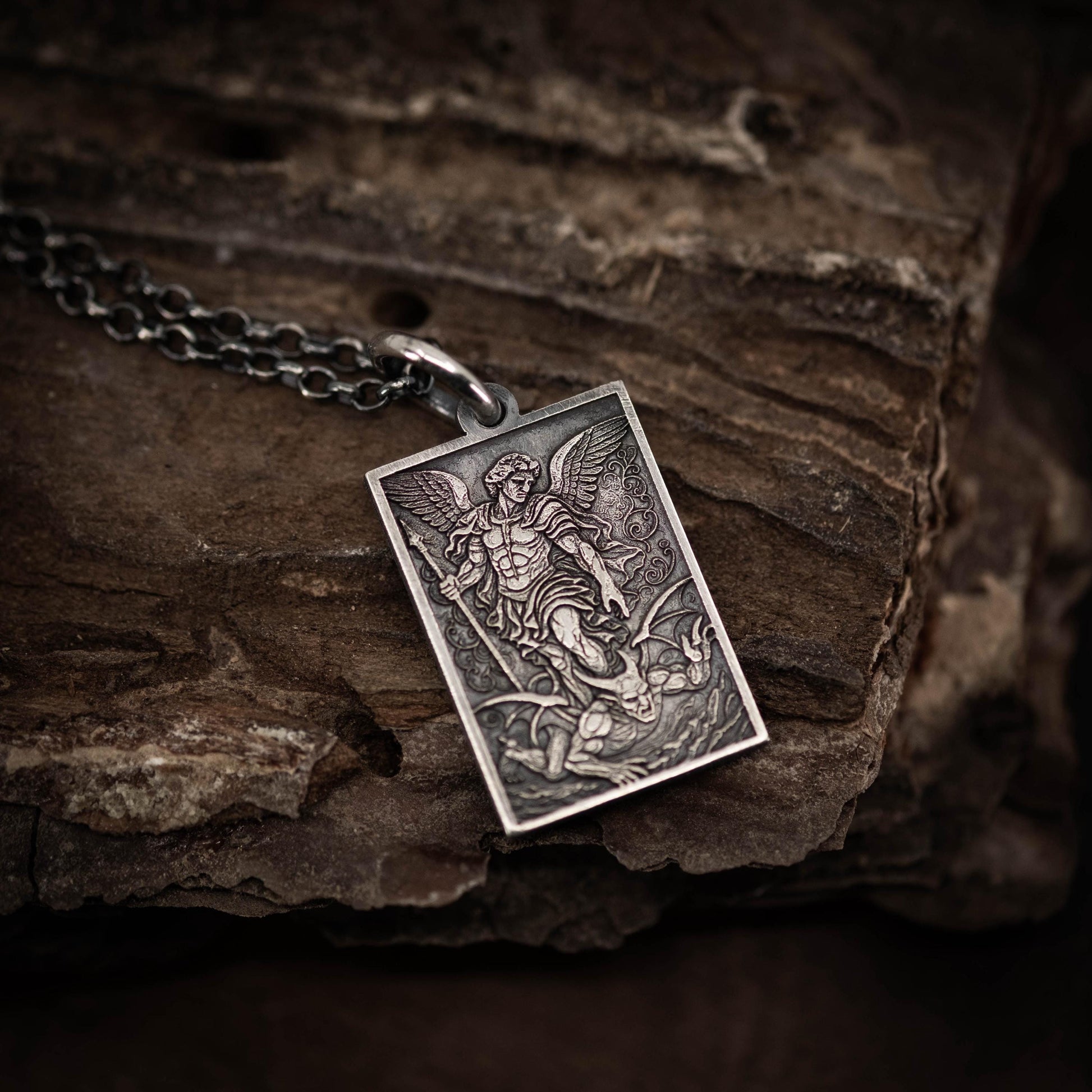 A silver pendant with an intricate design depicting a winged figure, possibly a saint or angel, is resting on a wooden surface.