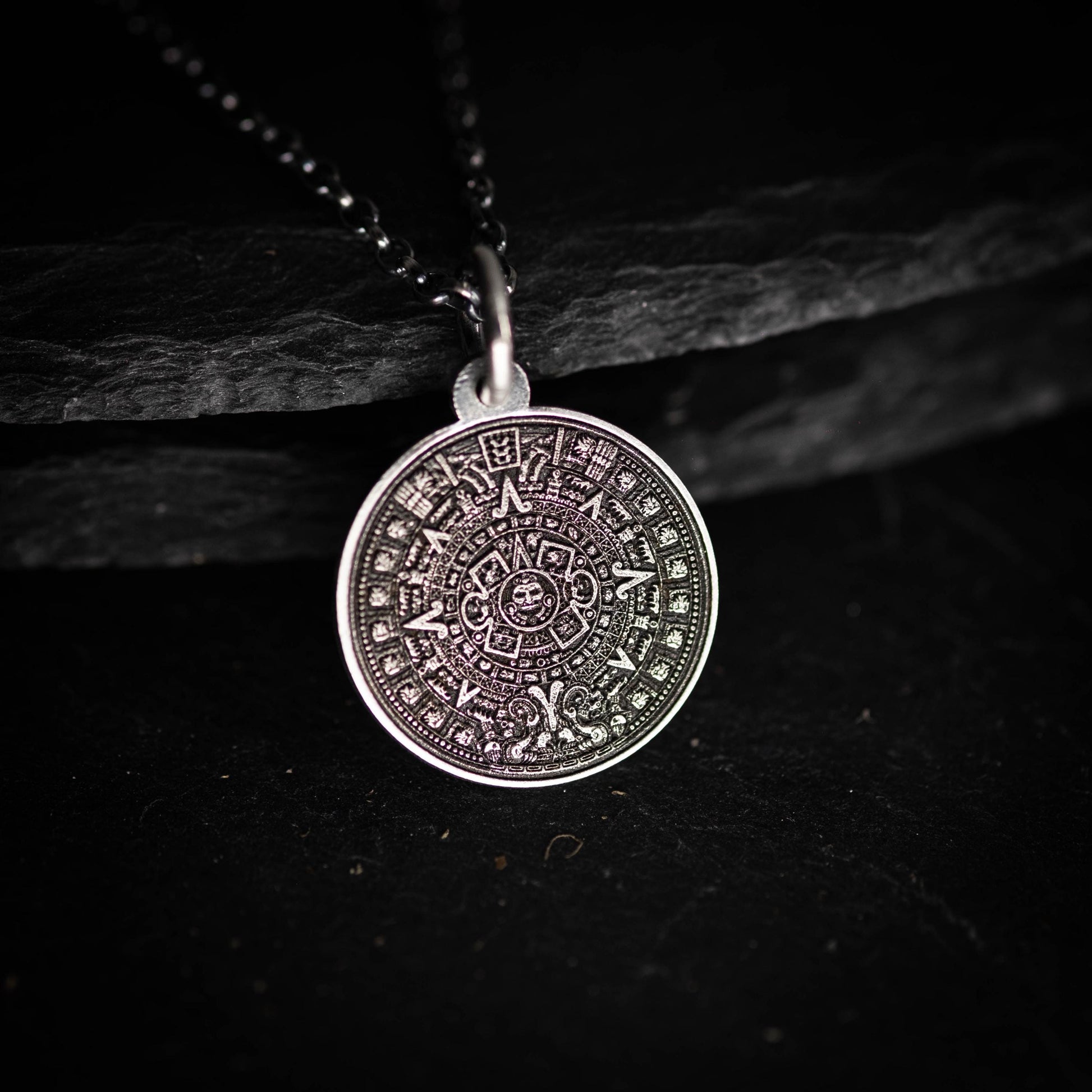 A silver pendant with an intricate design, resembling a calendar or calendar-like symbol, is displayed against a dark background.