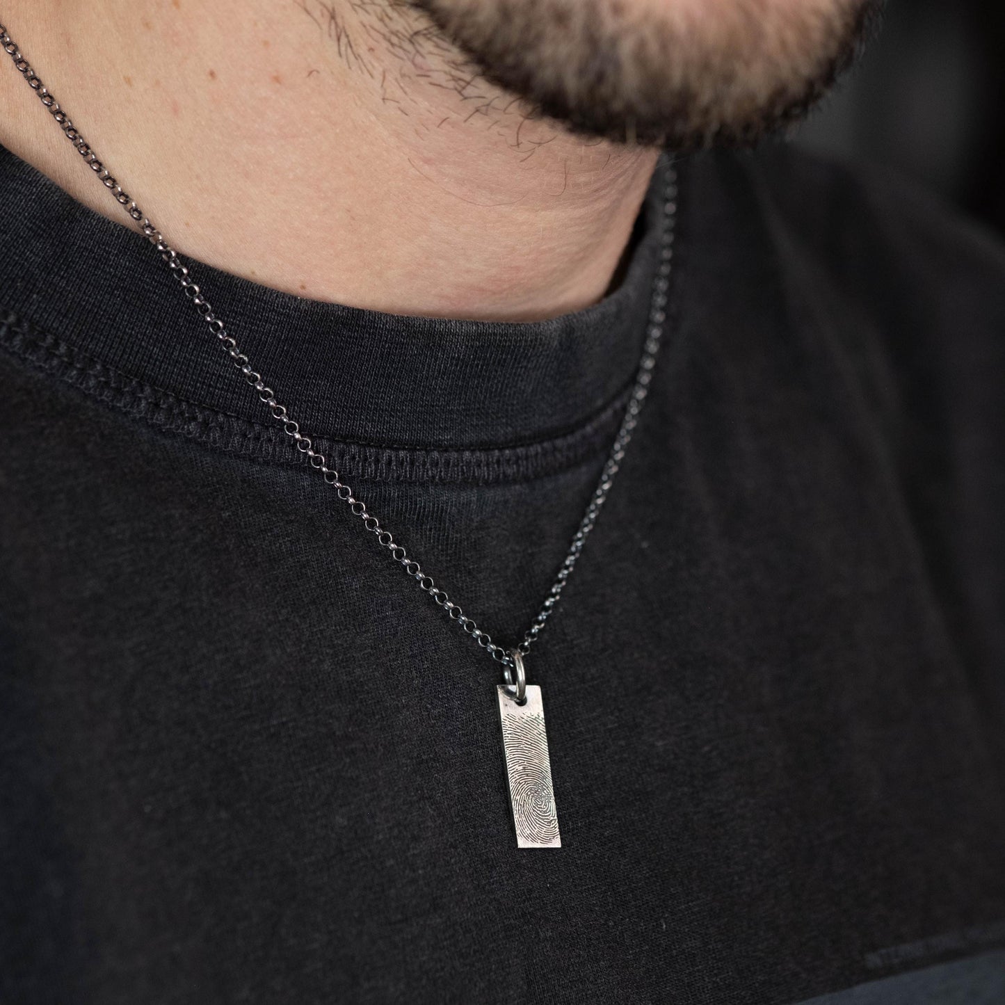 A close-up view of a person wearing a black t-shirt and a silver necklace with a rectangular pendant.
