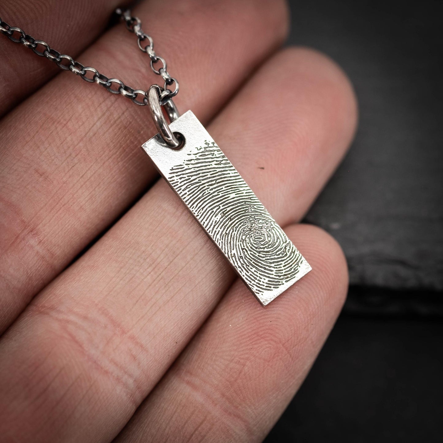 A hand holding a silver necklace with a rectangular pendant featuring a fingerprint design.