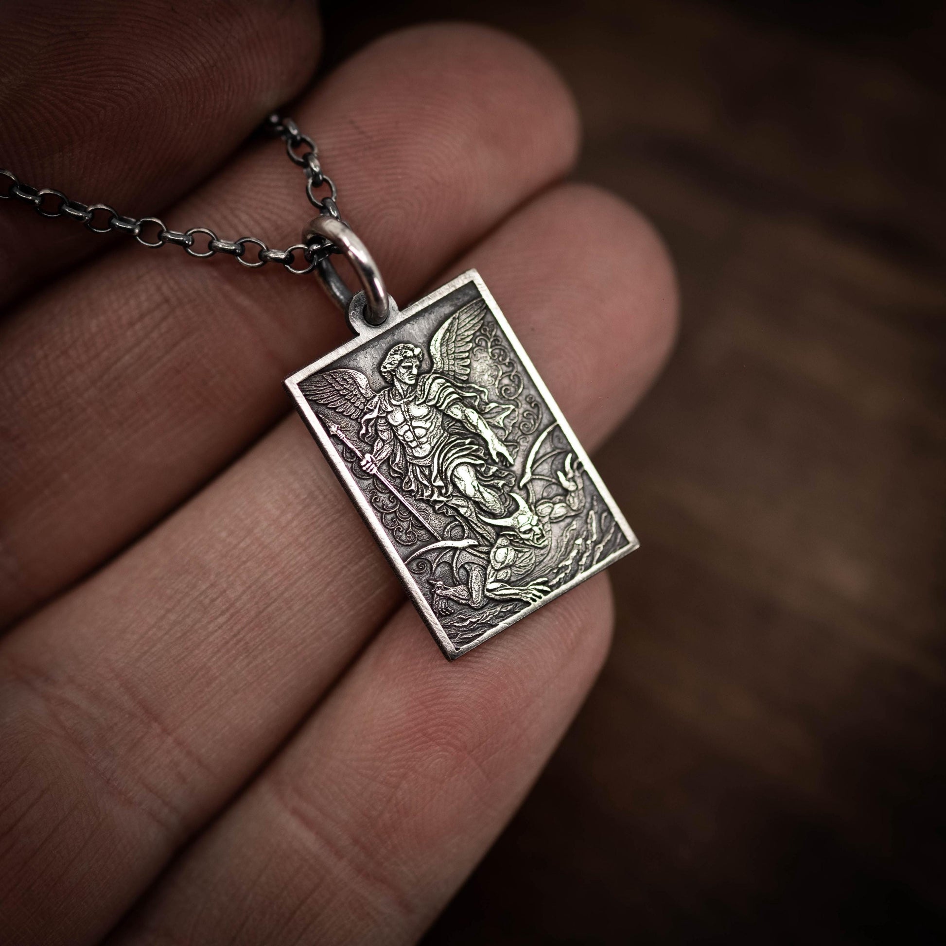 A hand holding a silver pendant with an engraved image of a winged figure, possibly a saint or angel, on a chain.
