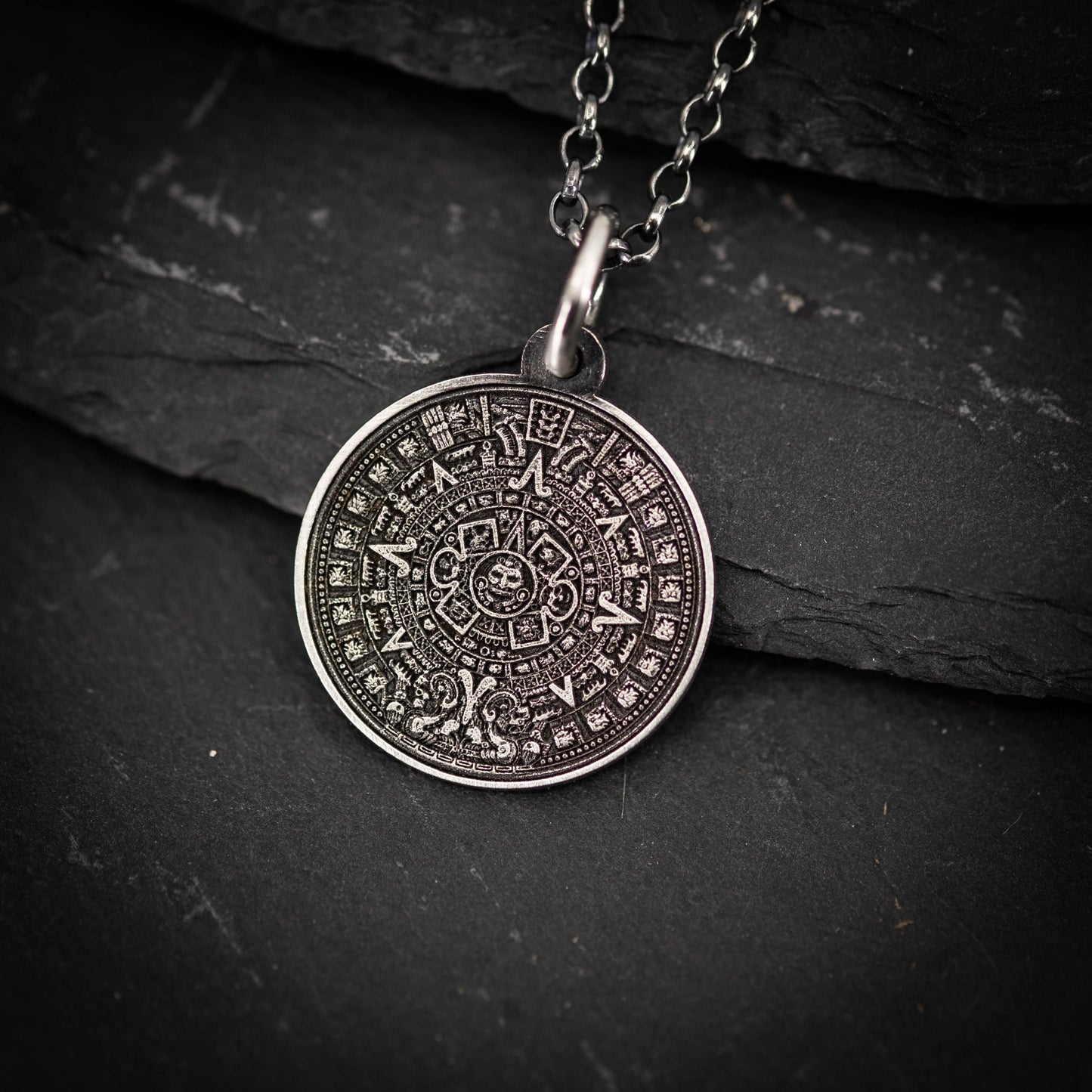 A silver pendant with an intricate design, resembling a calendar or calendar-like symbol, is displayed against a dark background.