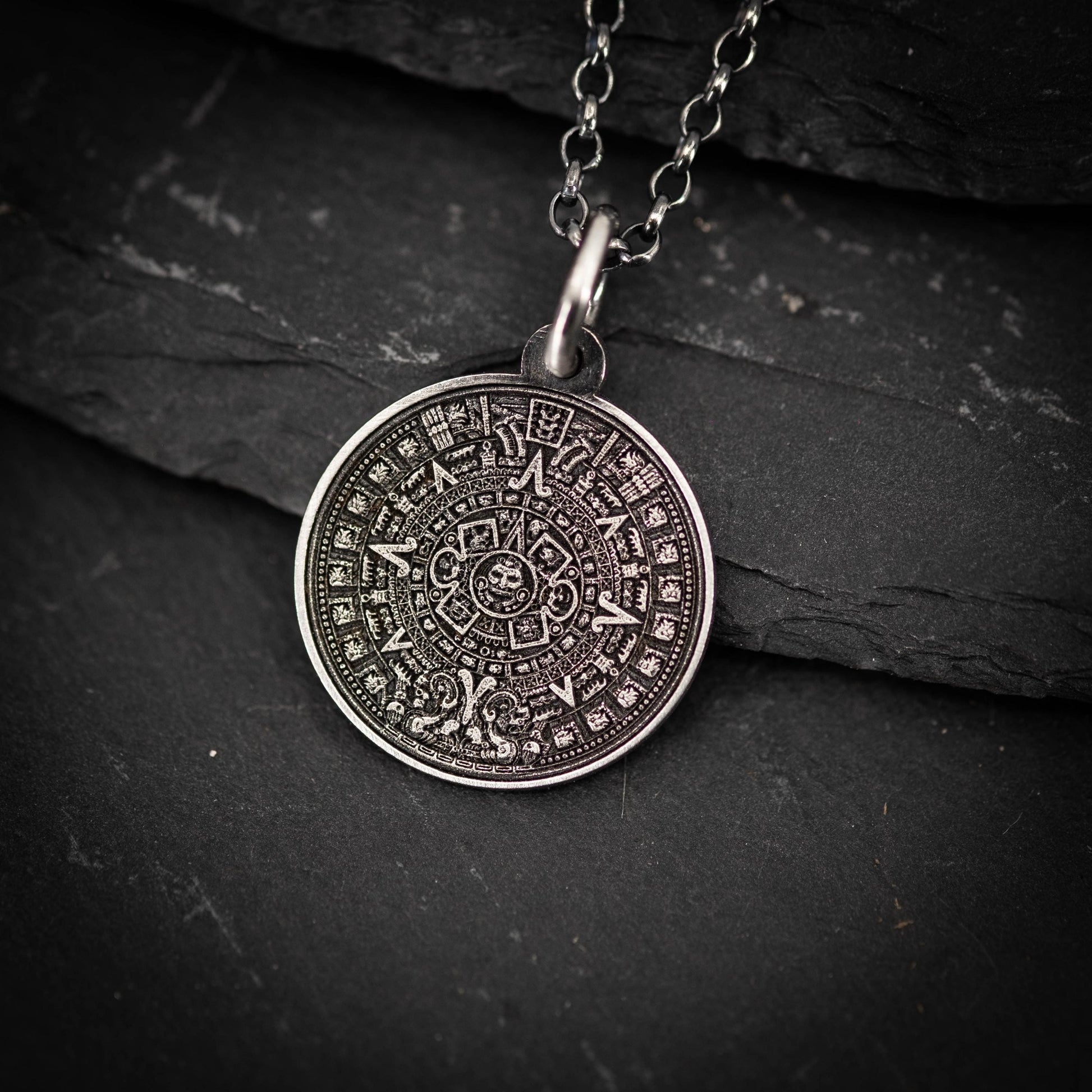A silver pendant with an intricate design, resembling a calendar or calendar-like symbol, is displayed against a dark background.