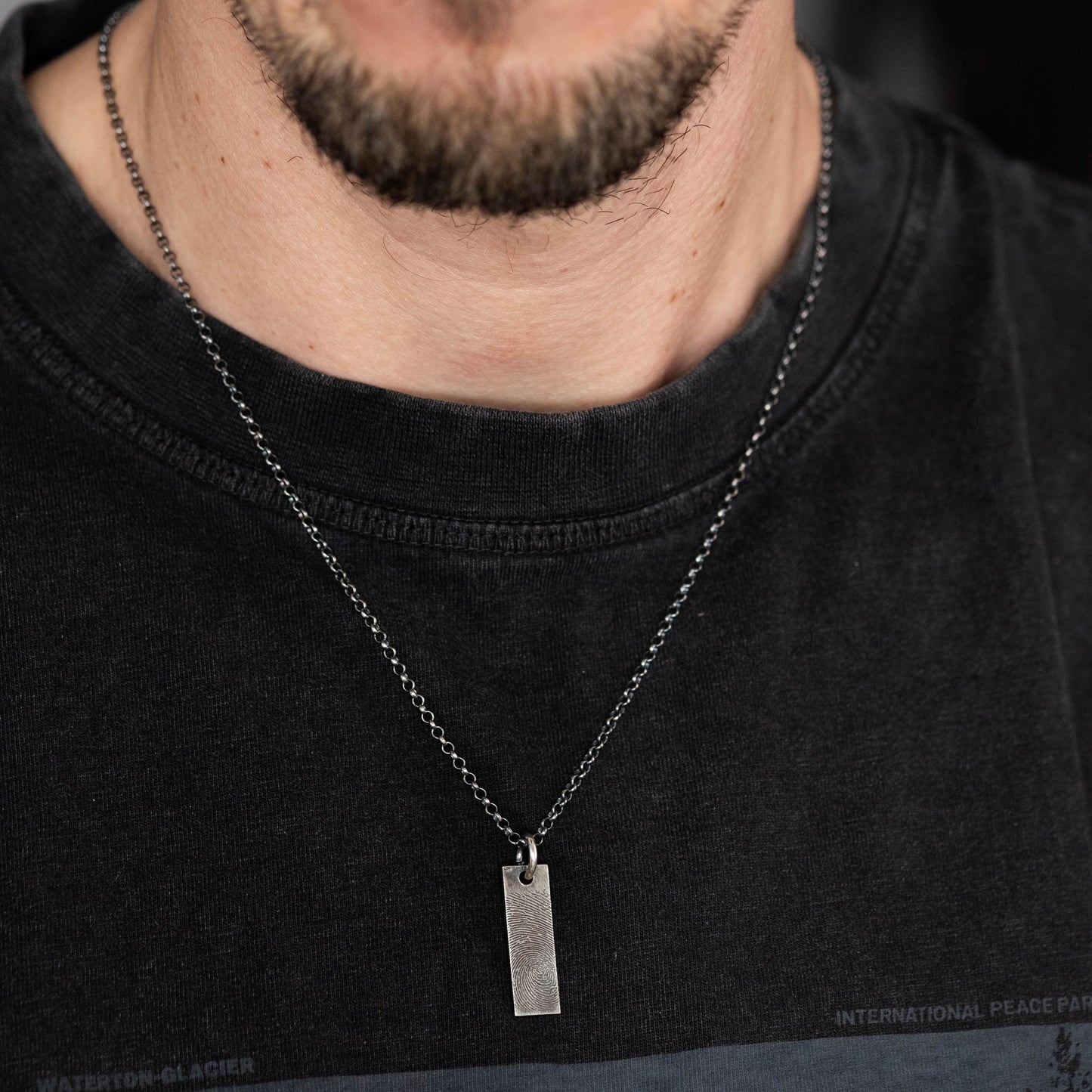 A close-up of a man wearing a black t-shirt and a silver necklace with a rectangular pendant.