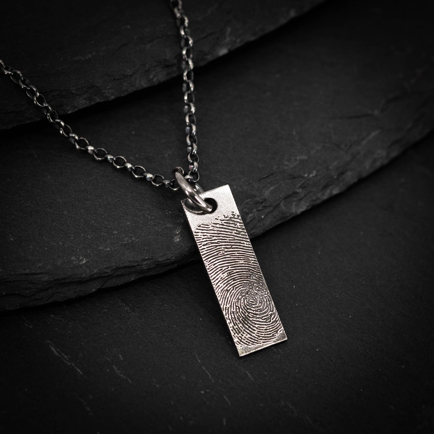 A silver necklace pendant with a fingerprint design is shown against a dark background.