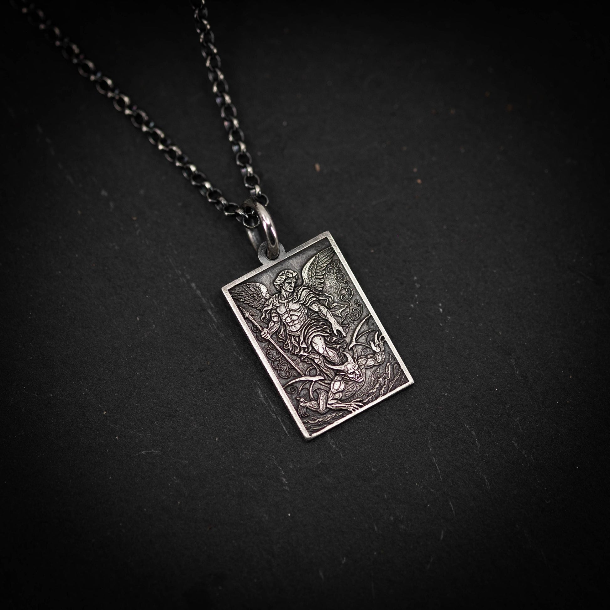 A rectangular pendant featuring an intricate design of a winged figure, possibly a saint or angel, against a dark background.