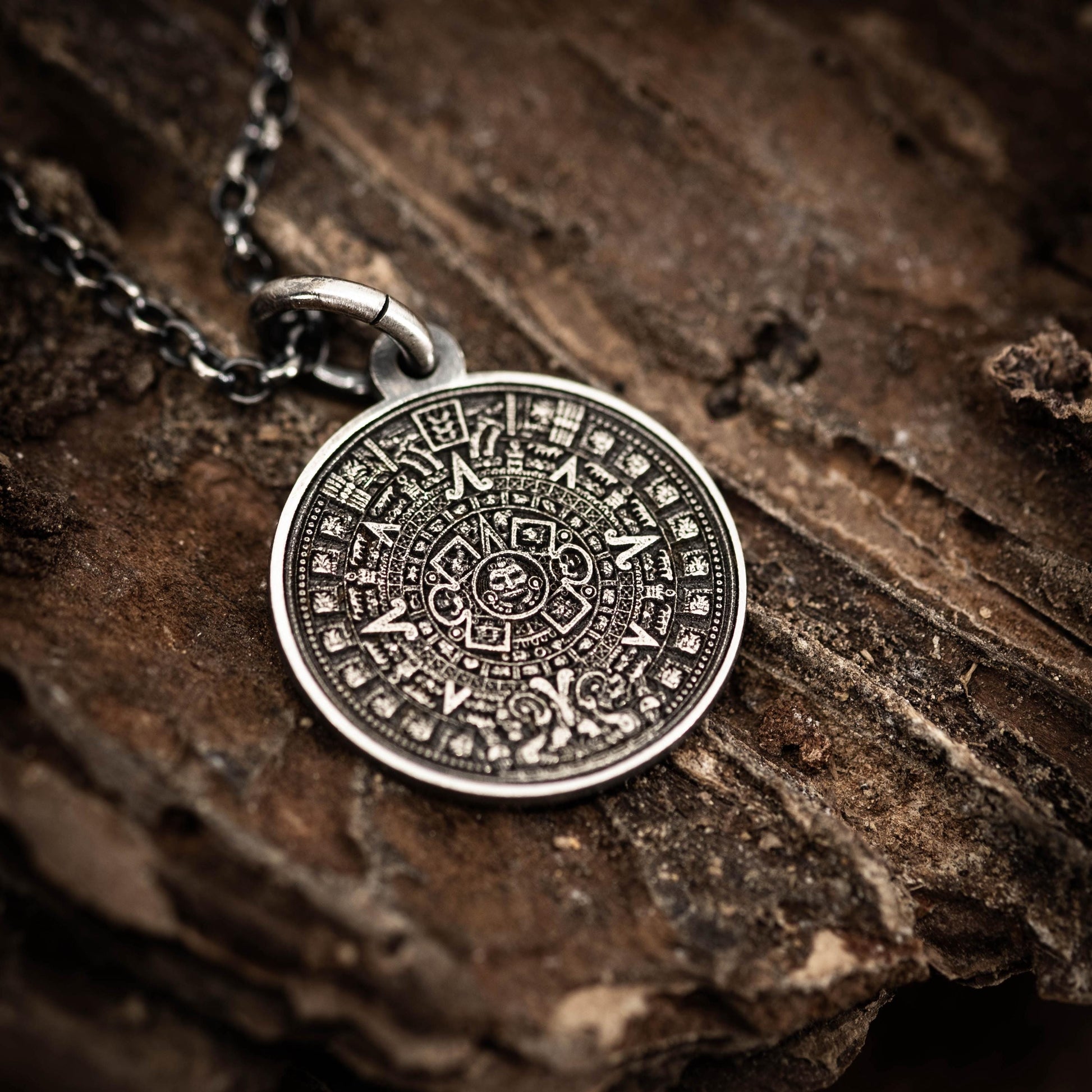A silver pendant with an intricate Mayan calendar design is displayed on a wooden surface.