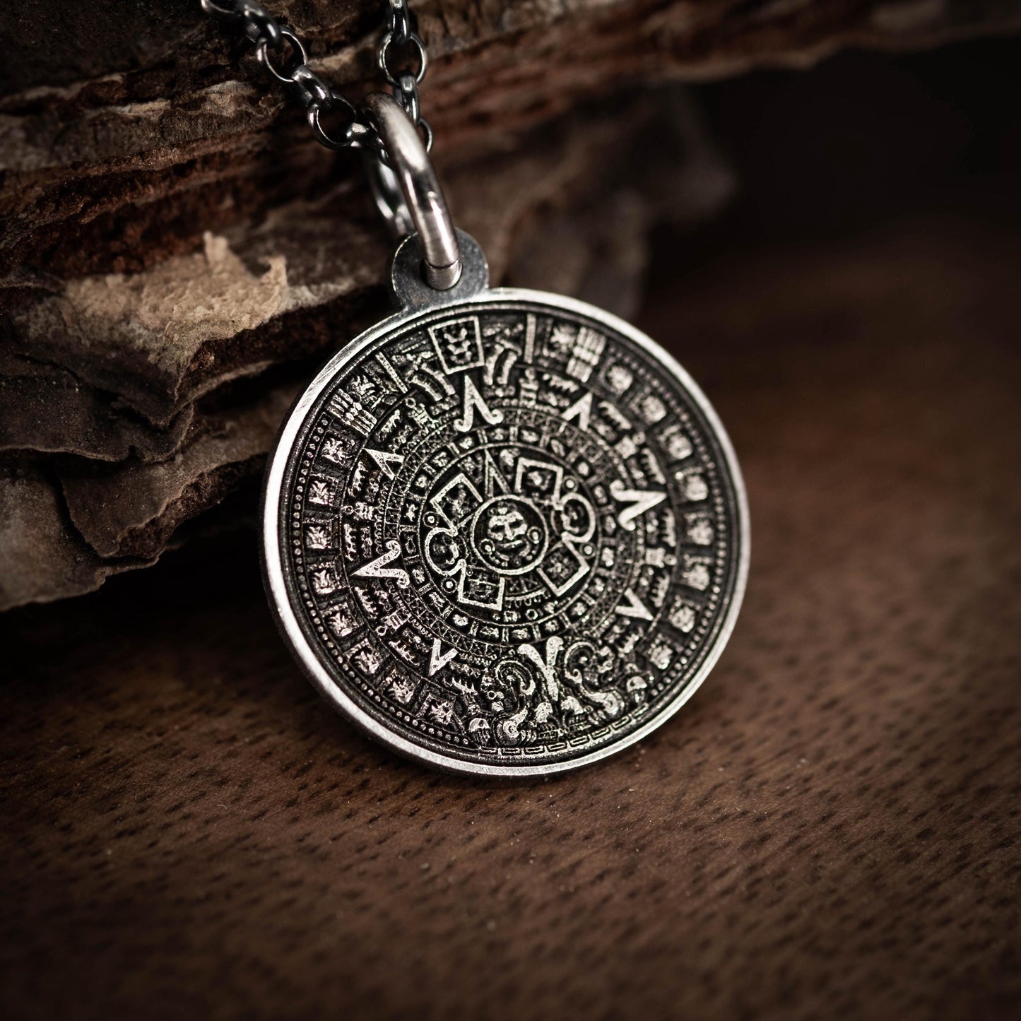 A silver pendant with an intricate Mayan calendar design is displayed on a wooden surface.