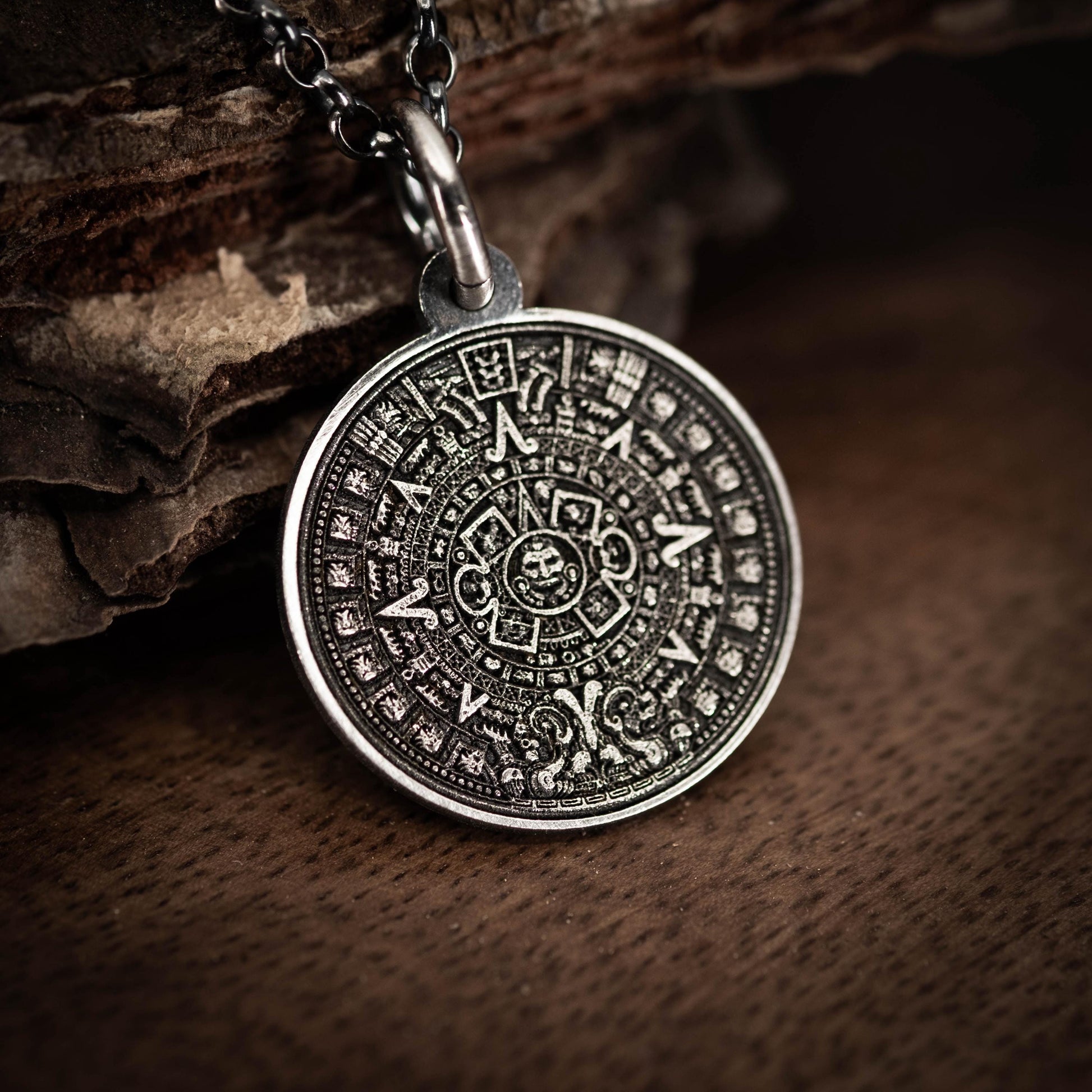 A silver pendant with an intricate Mayan calendar design is displayed on a wooden surface.