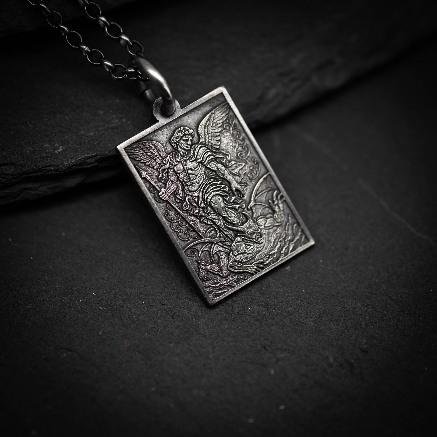 A silver pendant with an intricate design depicting a winged figure, possibly a saint or angel, on a dark surface.