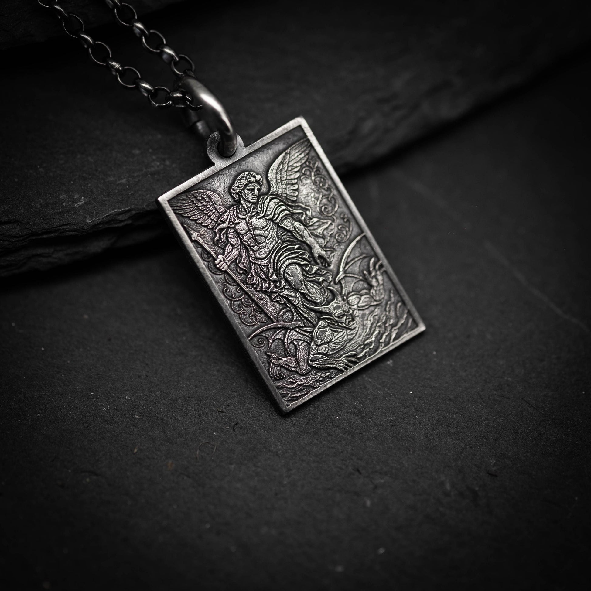 A silver pendant with an intricate design depicting a winged figure, possibly a saint or angel, on a dark surface.