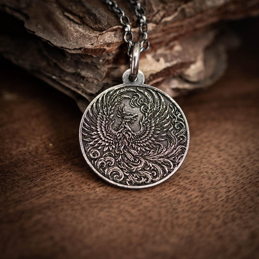 A silver pendant with an intricate design of a phoenix, a mythical bird, is displayed on a wooden surface.