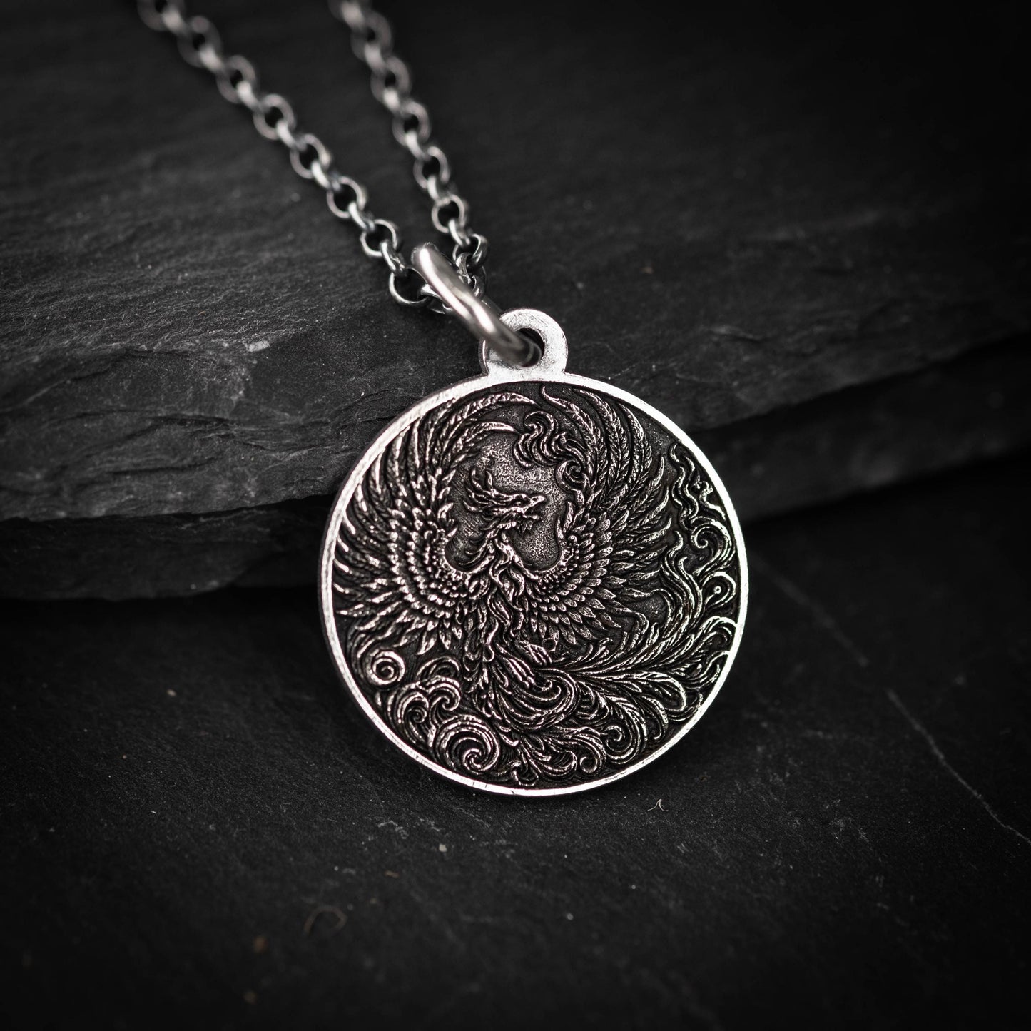 A silver pendant with an intricate design of a phoenix, a mythical bird, is displayed against a dark background.