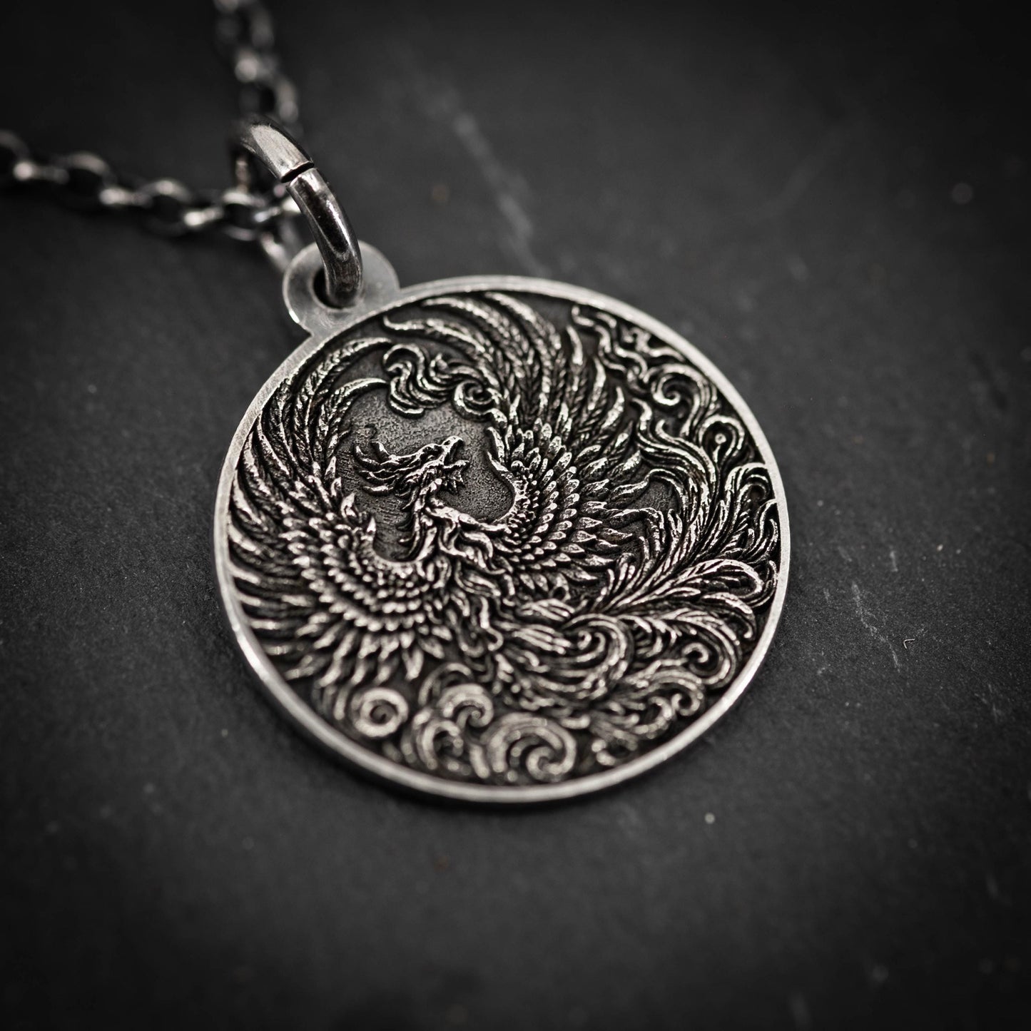 A close-up image of a silver pendant featuring an intricate design of a phoenix, a mythical bird that rises from the ashes.