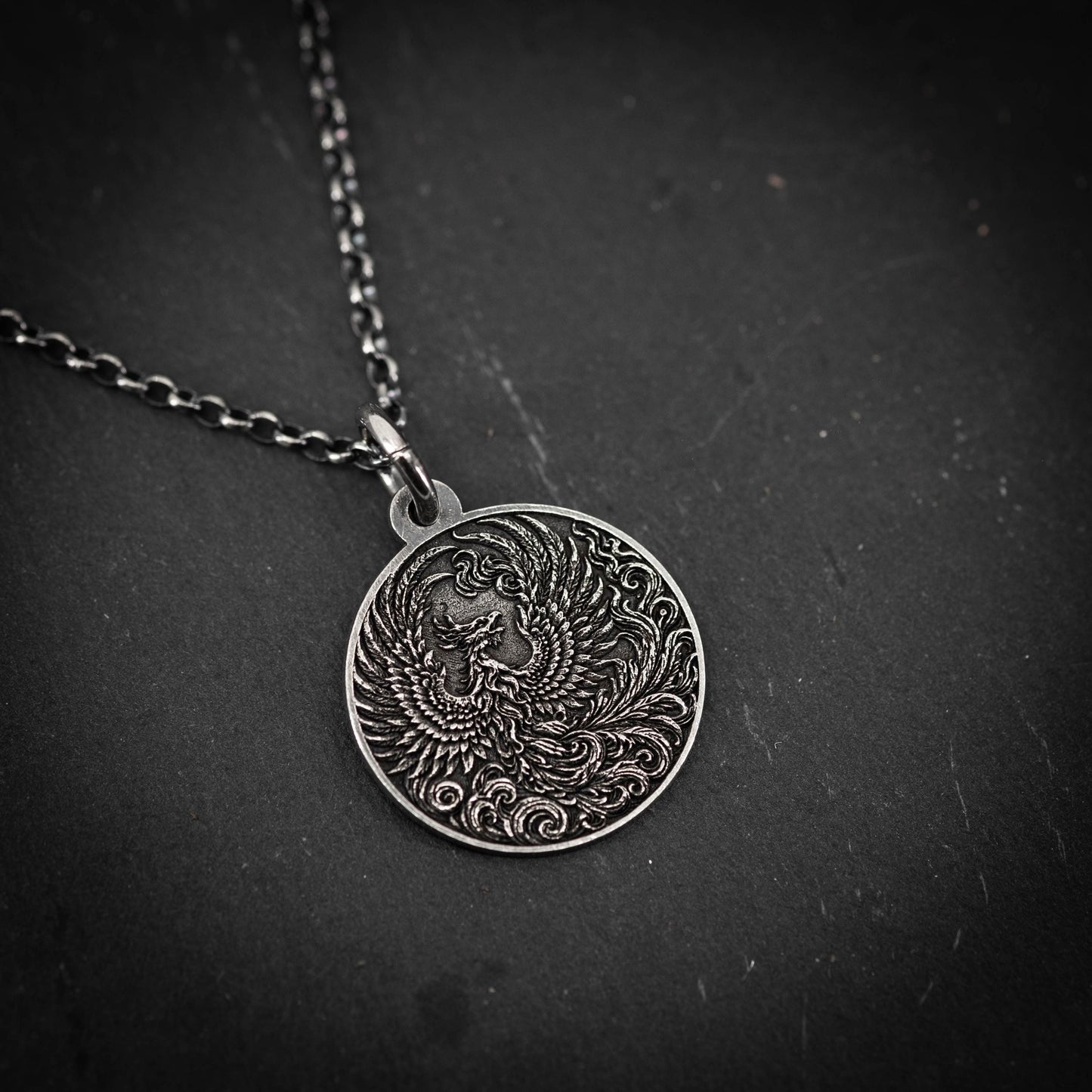 A silver pendant necklace with an intricate design of a phoenix, a mythical bird, on a dark surface.