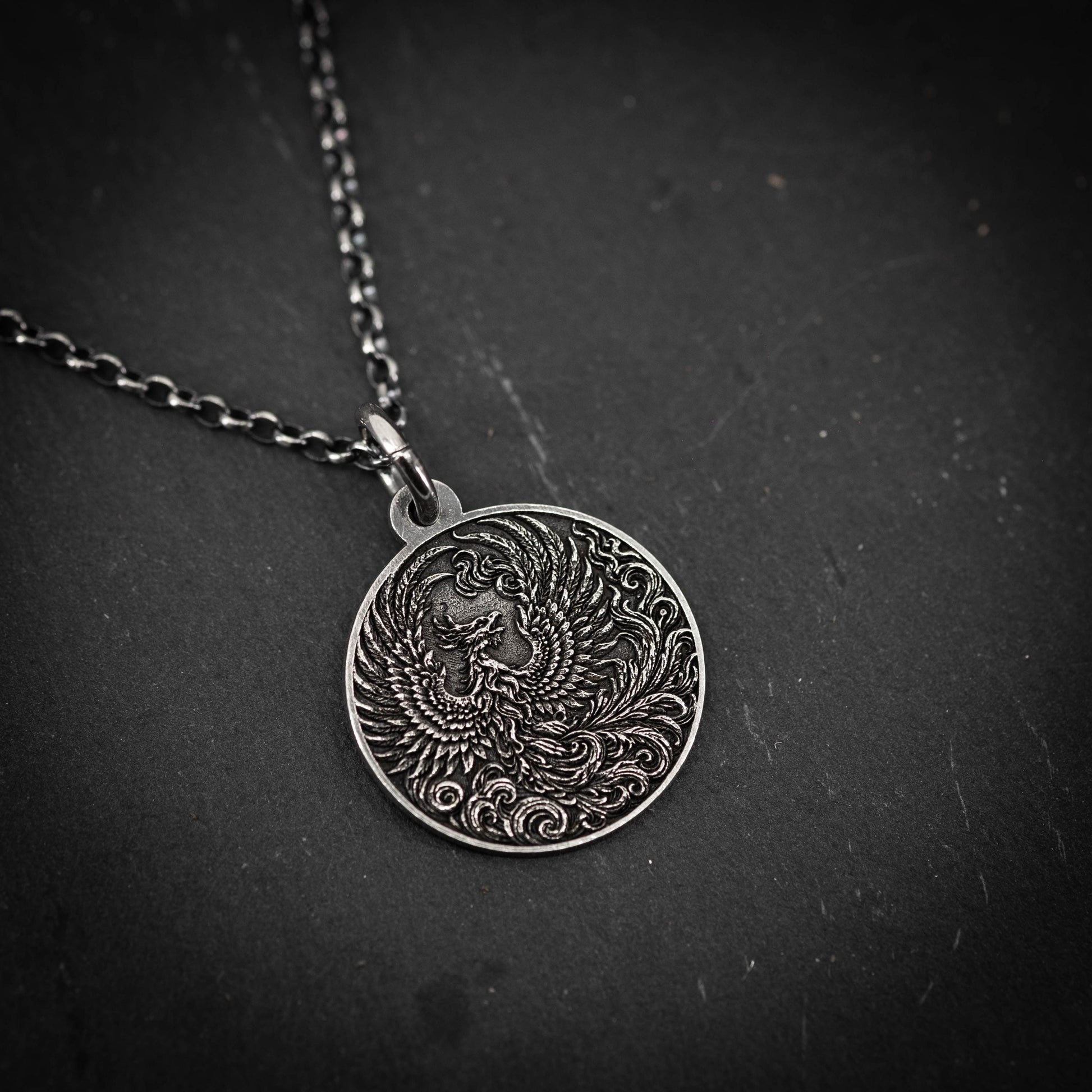 A silver pendant necklace with an intricate design of a phoenix, a mythical bird, on a dark surface.