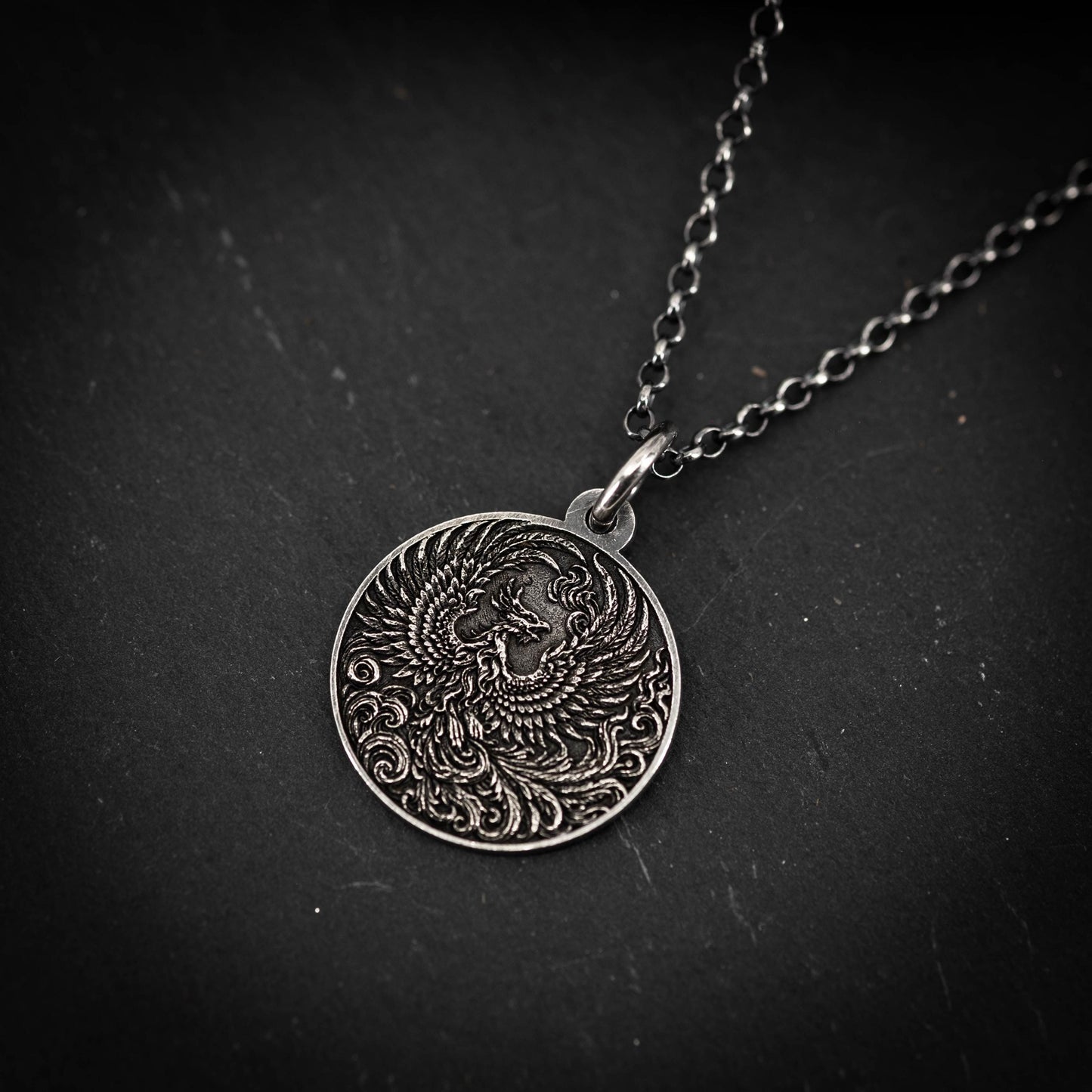 A silver pendant with an intricate design, featuring a central motif surrounded by swirling patterns, is displayed against a dark background.