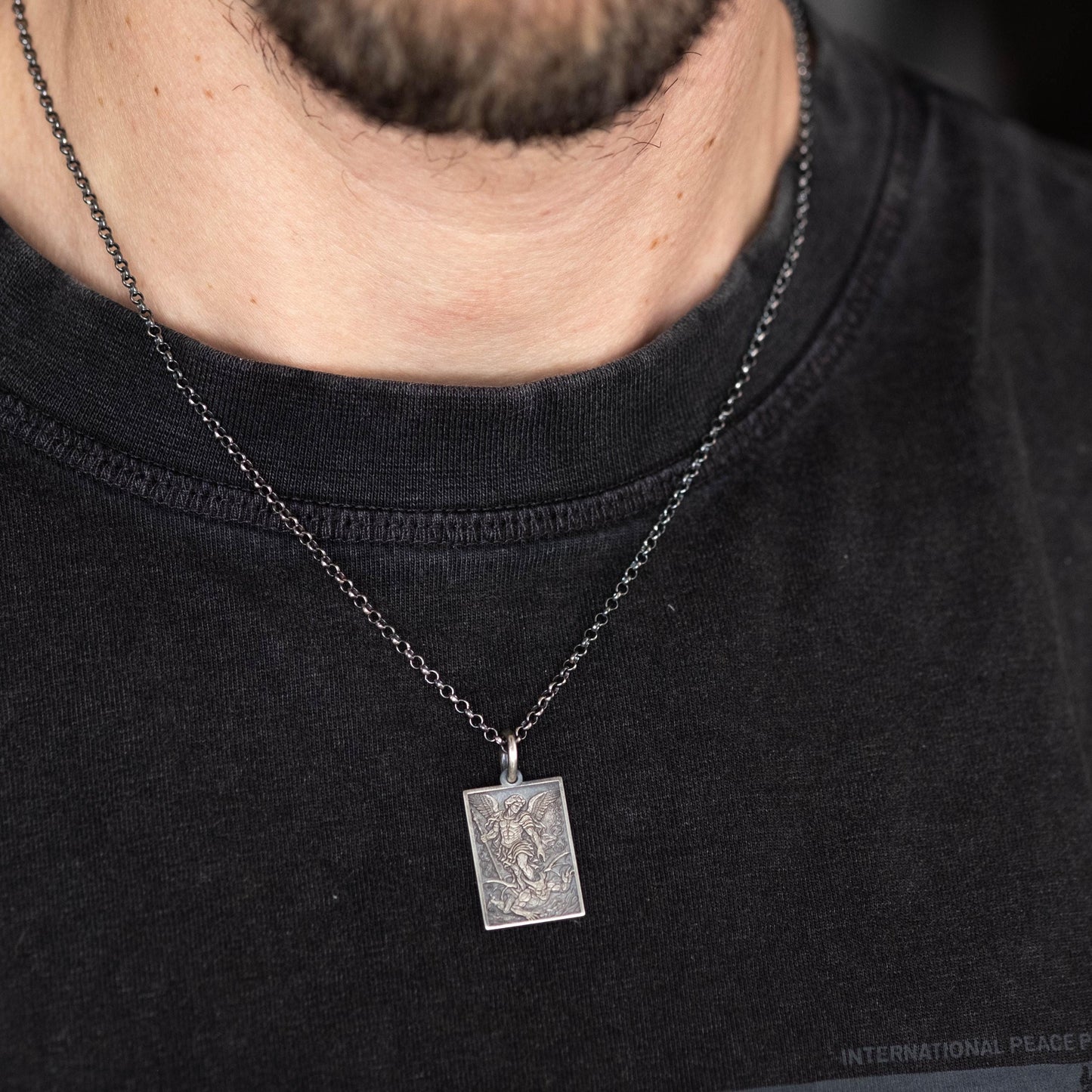 A close-up of a person wearing a black t-shirt with a silver pendant necklace featuring a small square pendant.