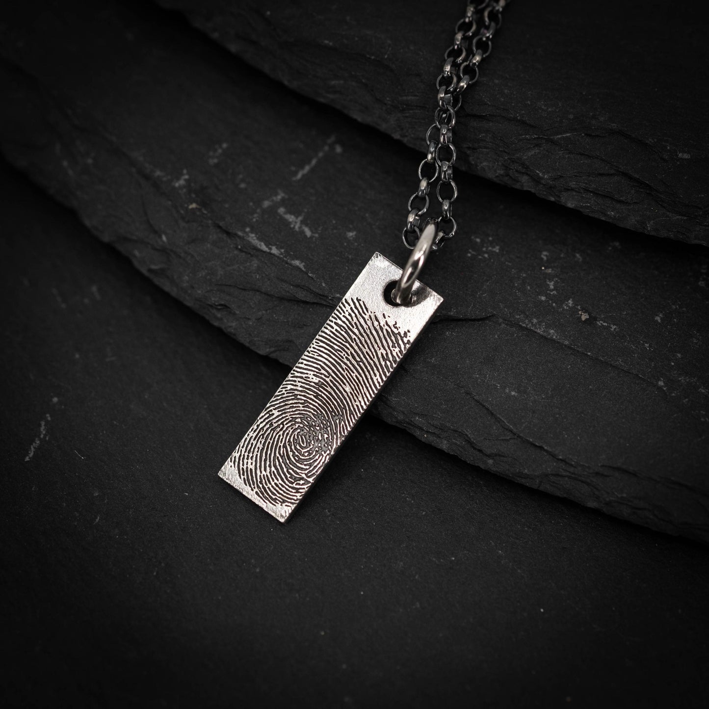 A silver necklace pendant with a fingerprint design is shown against a dark background.