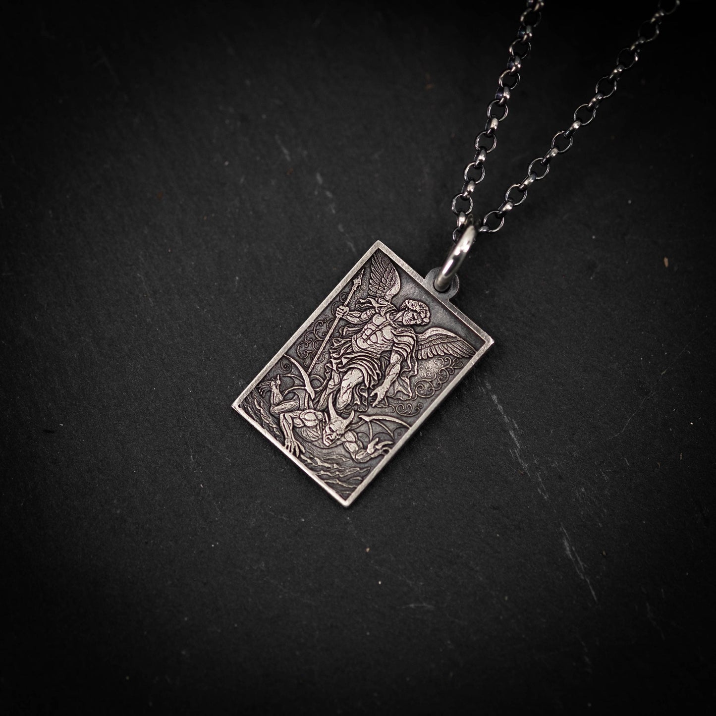 A rectangular pendant with an intricate design, featuring a figure with wings and a sword, is displayed against a dark background.