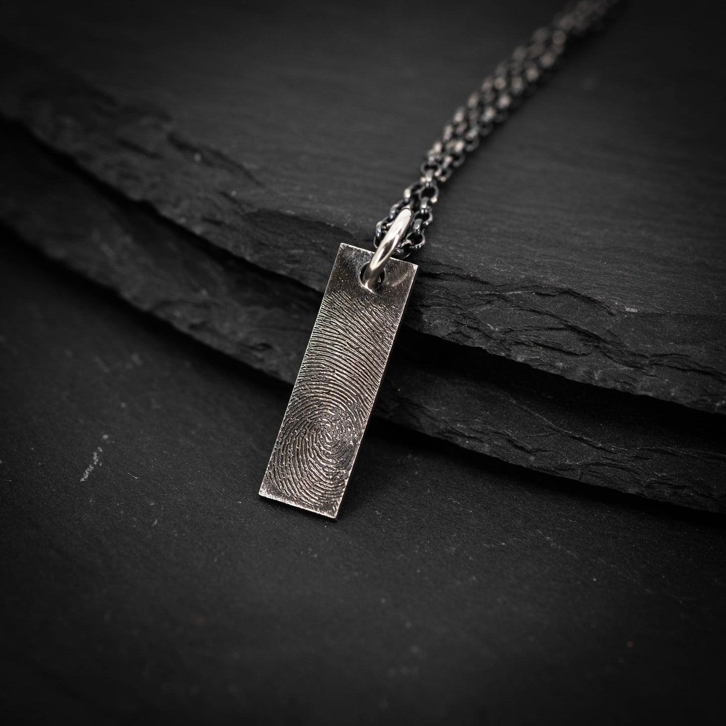 A silver pendant with a fingerprint design is shown against a dark background.