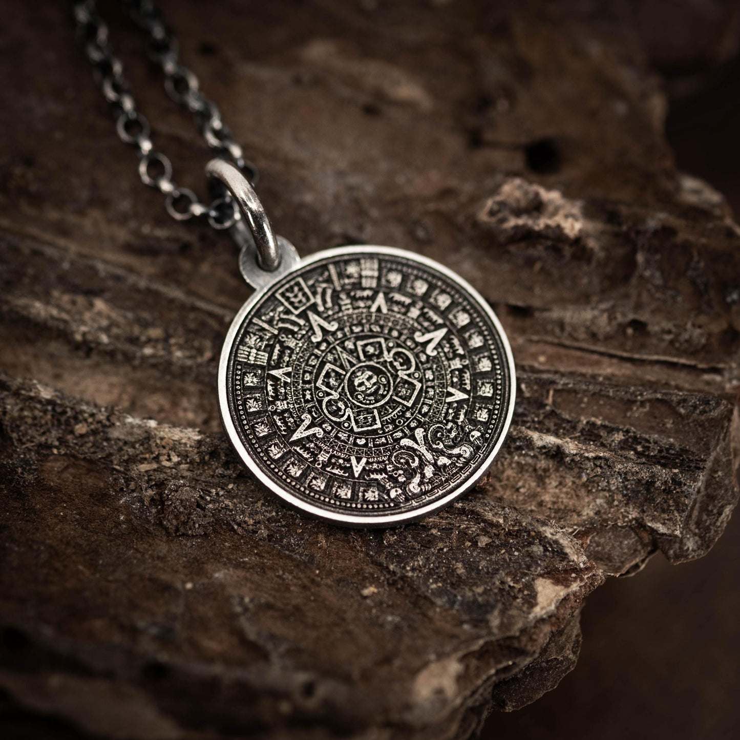 A silver pendant with an intricate design, resembling a calendar or calendar-like symbol, is displayed on a rough, textured surface.