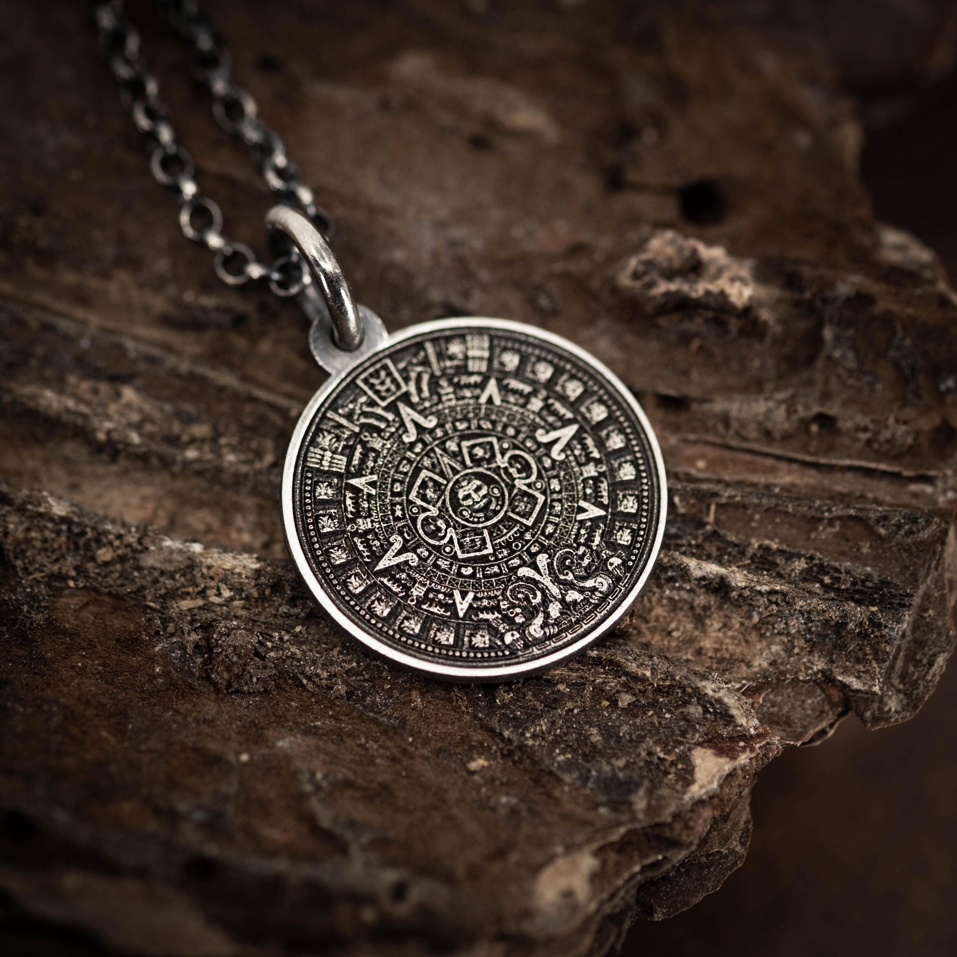 A silver pendant with an intricate design, resembling a calendar or calendar-like symbol, is displayed on a rough, textured surface.