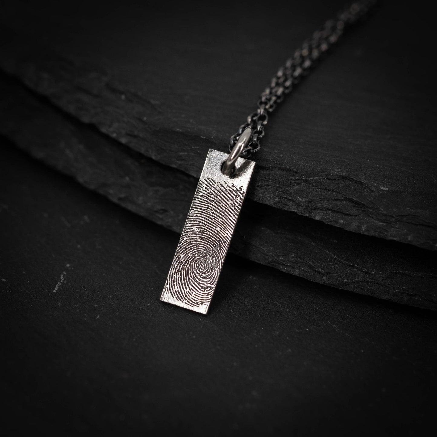 A silver necklace pendant with a fingerprint design rests on a dark surface.