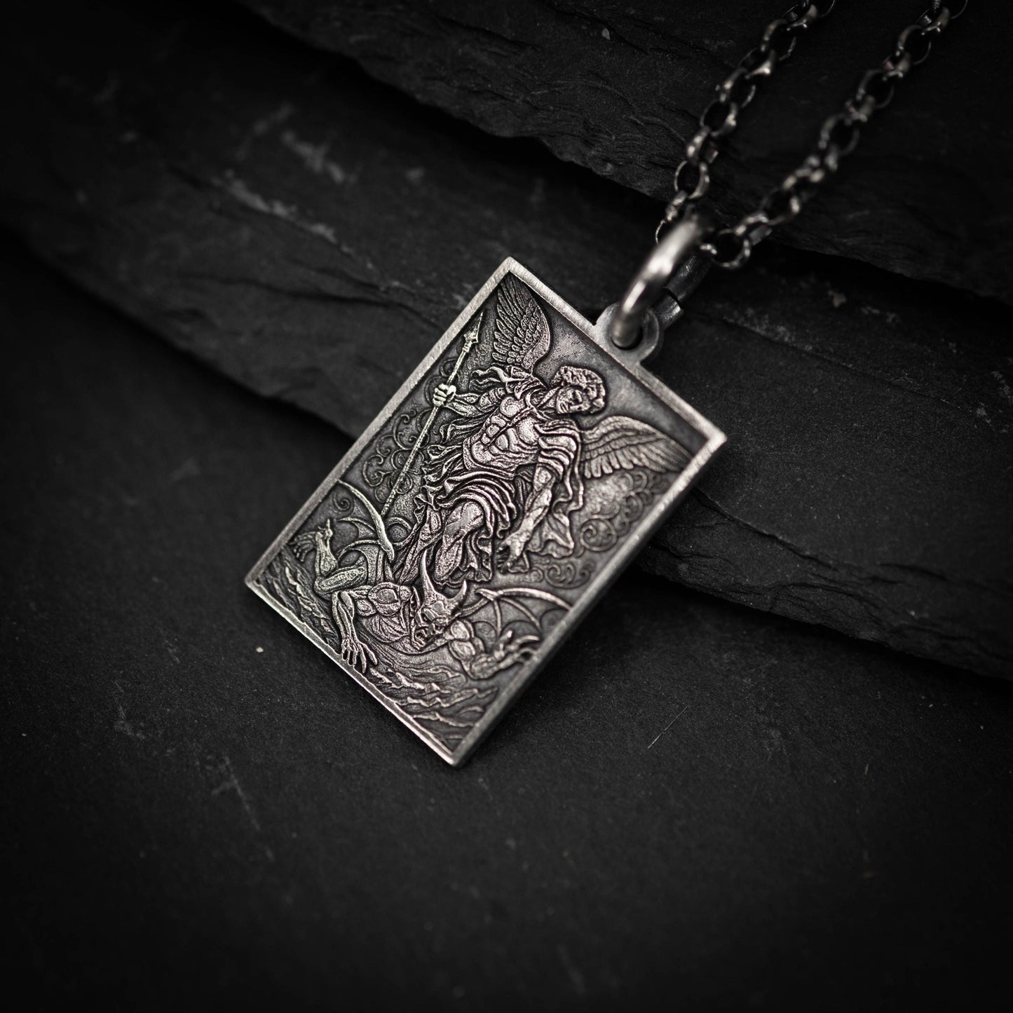 A silver pendant with an intricate design depicting a winged figure, possibly a deity or angel, on a dark surface.