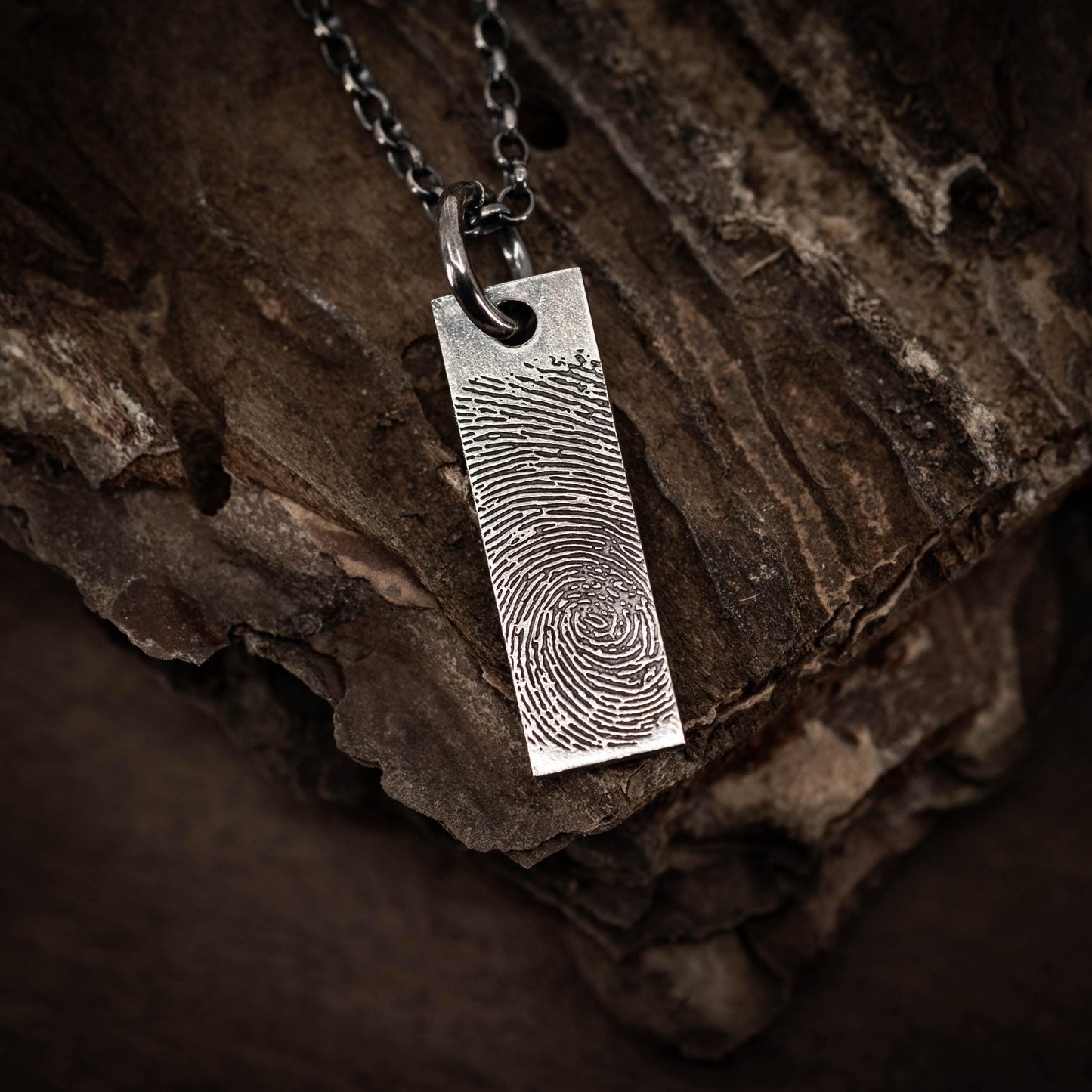A silver pendant with a fingerprint design is hanging from a chain against a dark, textured background.