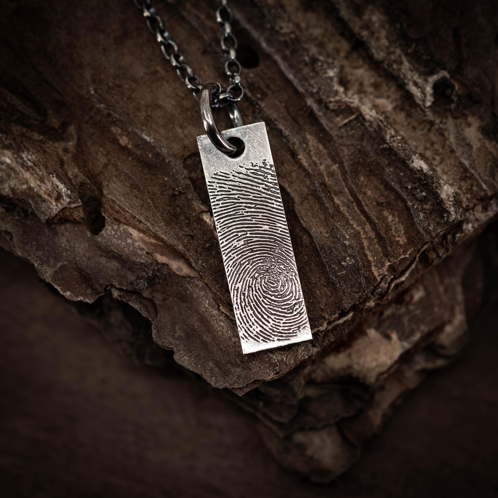 A silver pendant with a fingerprint design is hanging from a chain against a dark, textured background.