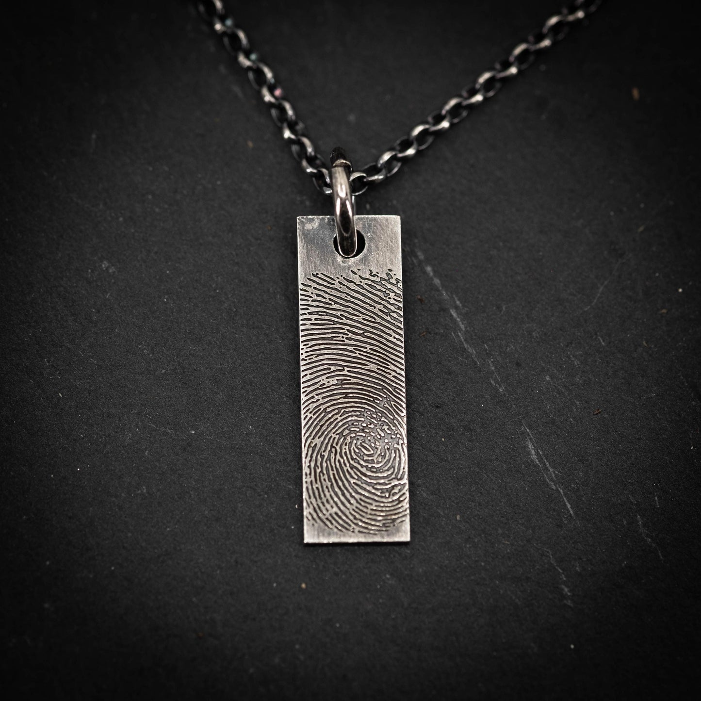 A silver rectangular pendant with a fingerprint design on it, suspended from a chain against a dark background.