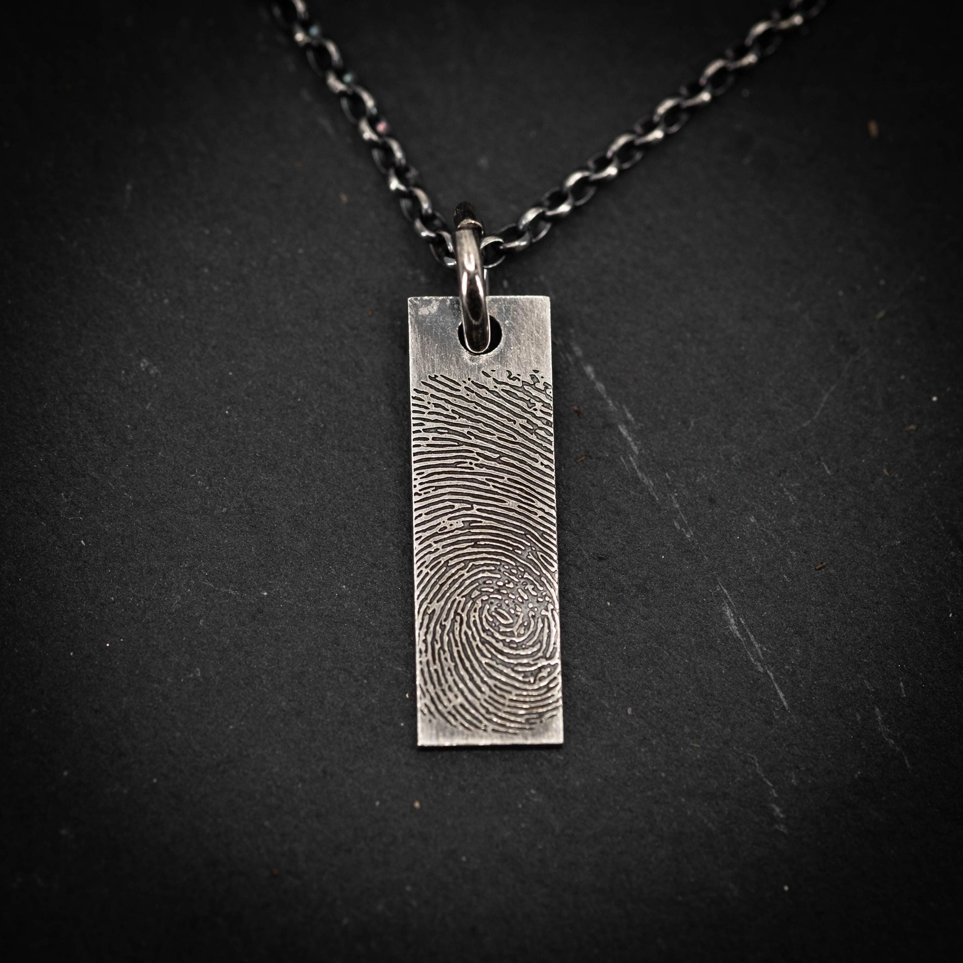 A silver rectangular pendant with a fingerprint design on it, suspended from a chain against a dark background.