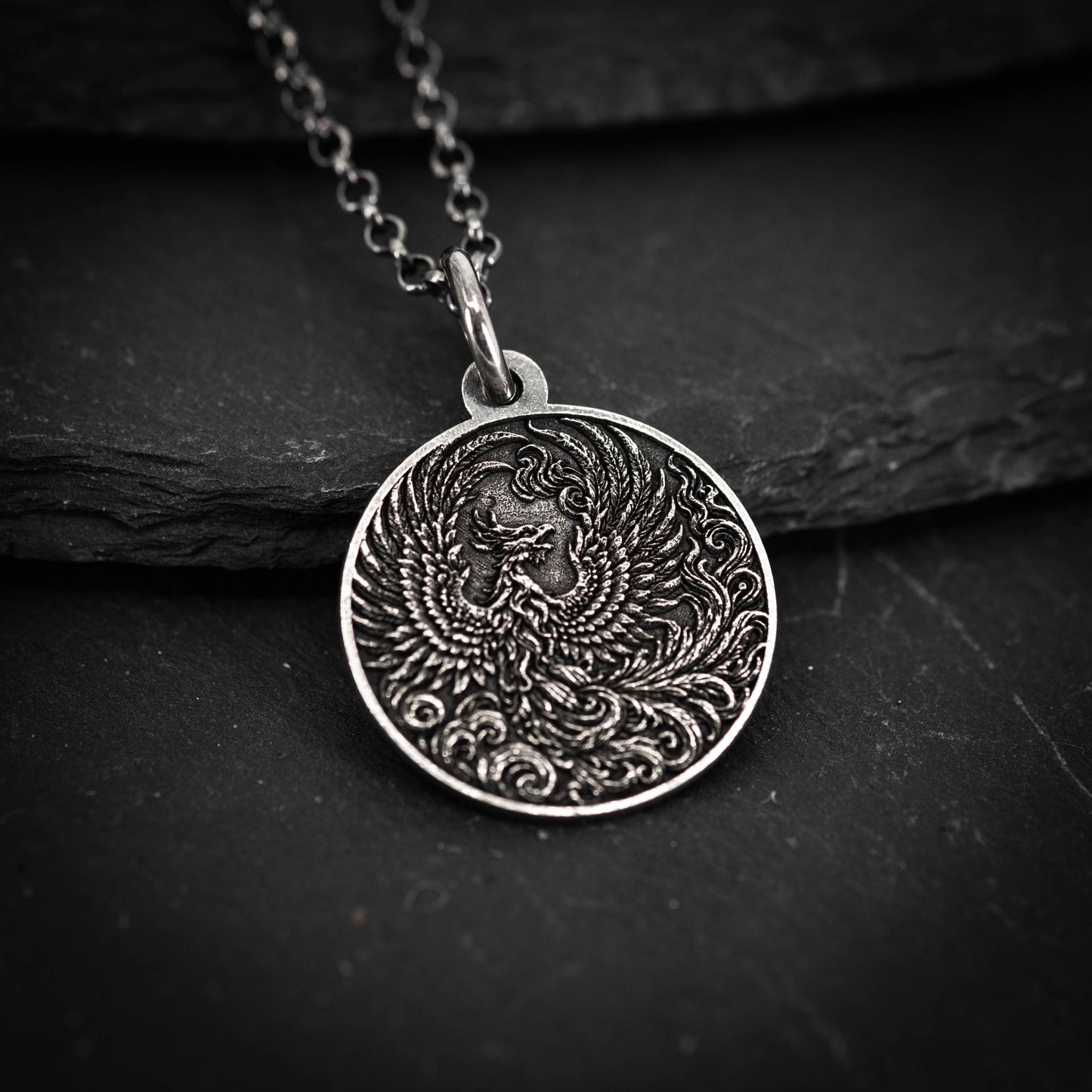 A silver pendant with an intricate design of a phoenix, a mythical bird, is displayed against a dark background.