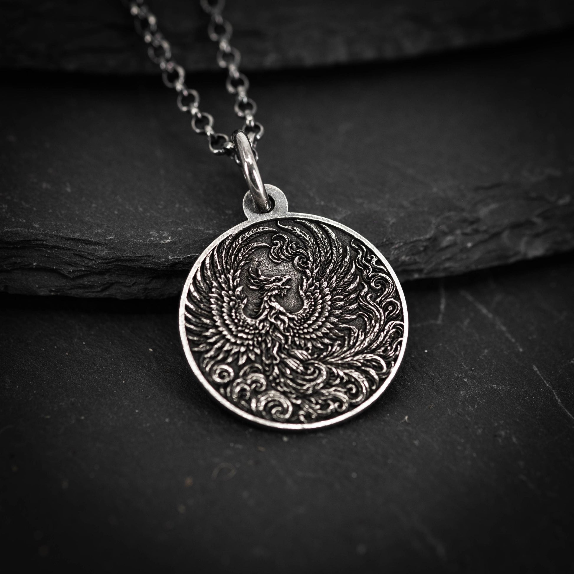 A silver pendant with an intricate design of a phoenix, a mythical bird, is displayed against a dark background.