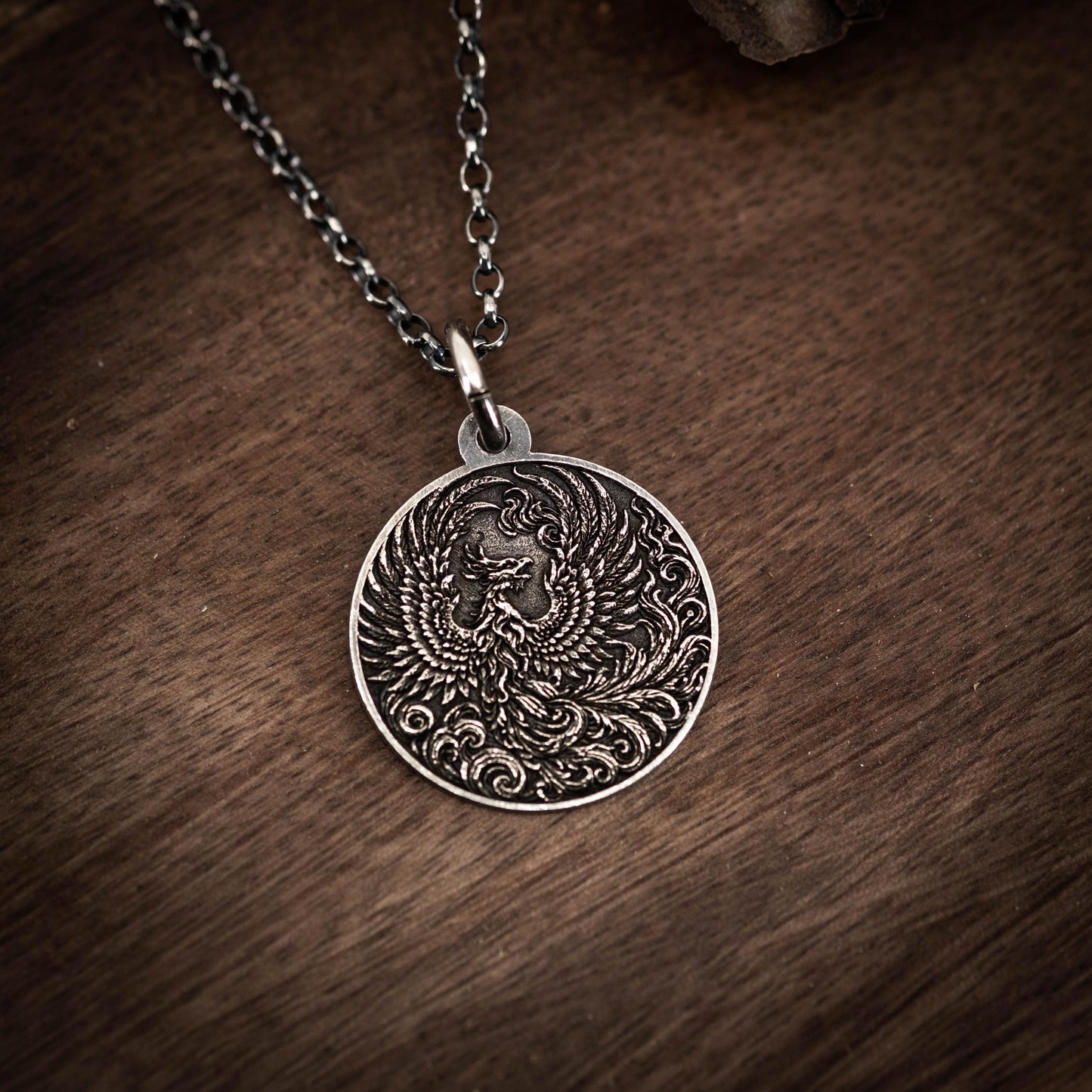 A silver pendant with an intricate design, featuring a bird-like creature with wings spread wide, is displayed on a wooden surface.