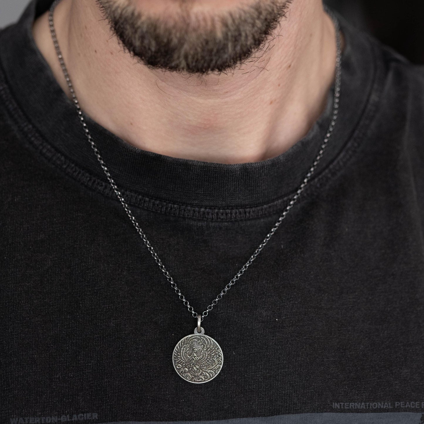 A close-up of a man wearing a black t-shirt and a silver necklace with a round pendant.