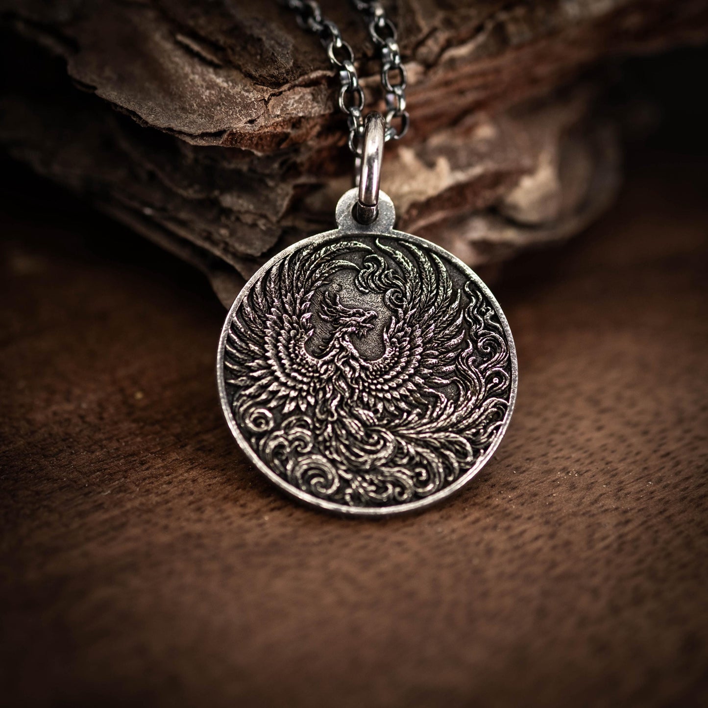 A silver pendant with an intricate design of a phoenix, a mythical bird, is displayed on a wooden surface.