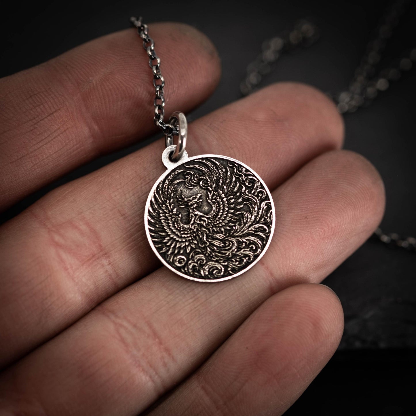 A person's hand holding a silver pendant with an intricate design, including a bird-like figure.