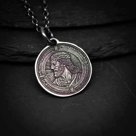 A silver-colored pendant featuring a detailed engraved image of a man's face, possibly depicting Jesus Christ.