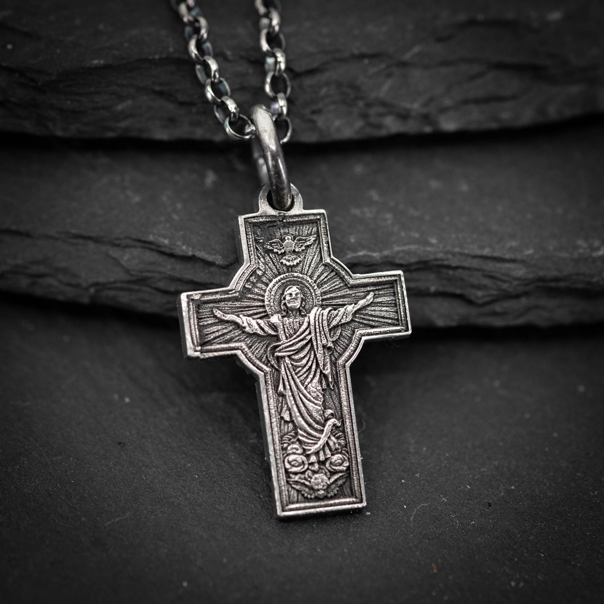 A silver cross pendant with a religious figure on it, hanging on a chain against a dark background.
