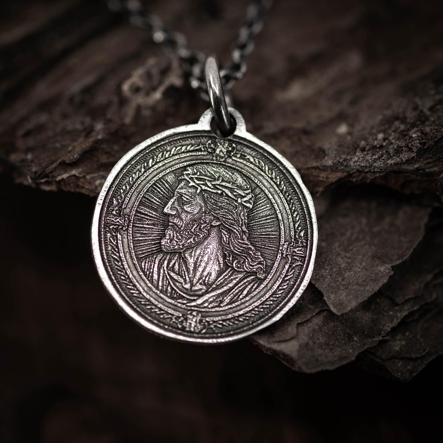 A silver-colored pendant featuring a detailed engraved image of a man's face, possibly a religious figure, is displayed on a chain against a dark background.