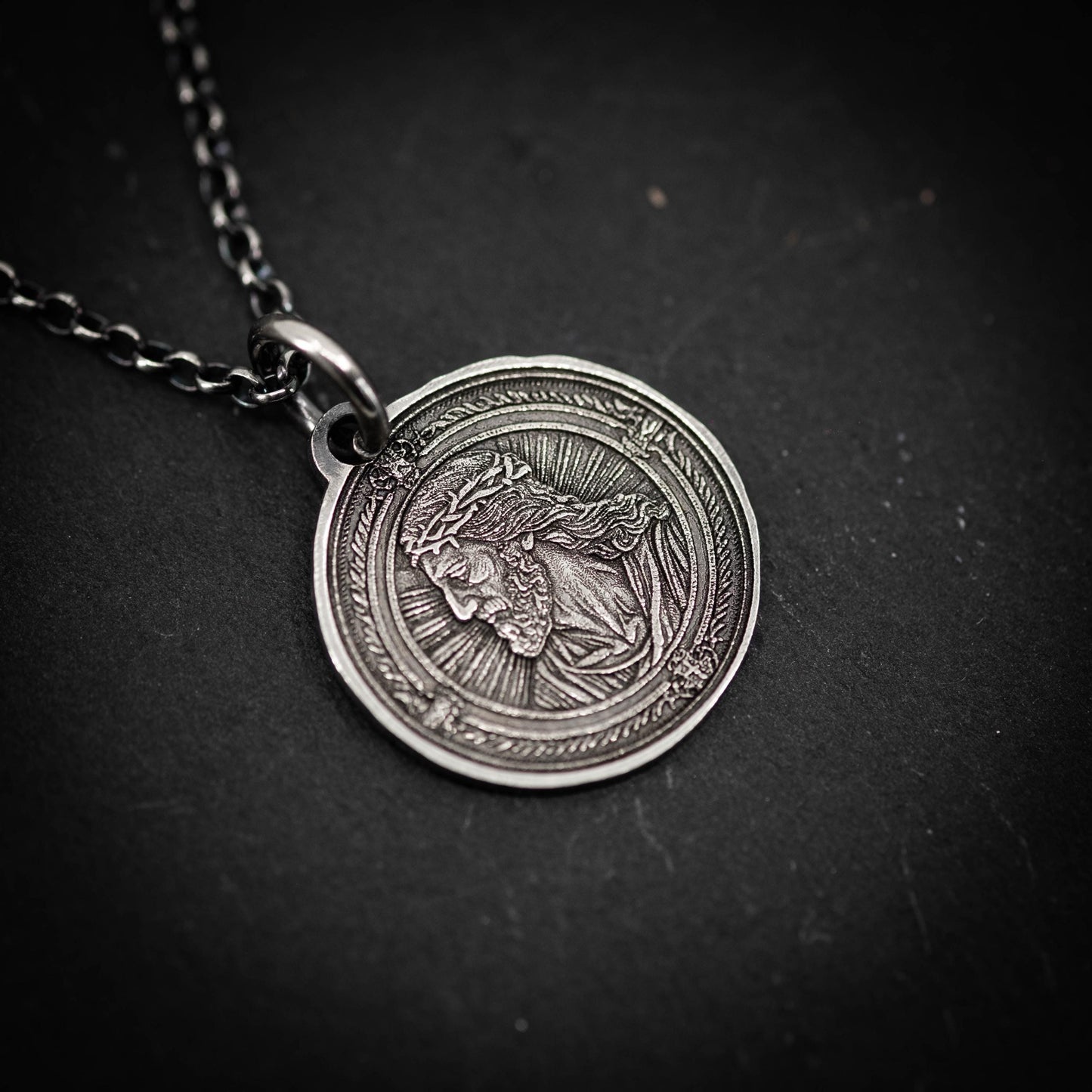 A silver-colored pendant featuring a detailed engraved image of a religious figure, likely a saint or deity, on a dark background.