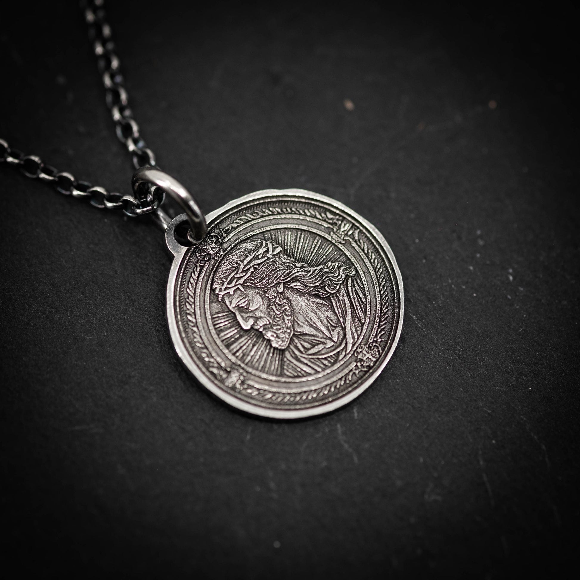 A silver-colored pendant featuring a detailed engraved image of a religious figure, likely a saint or deity, on a dark background.