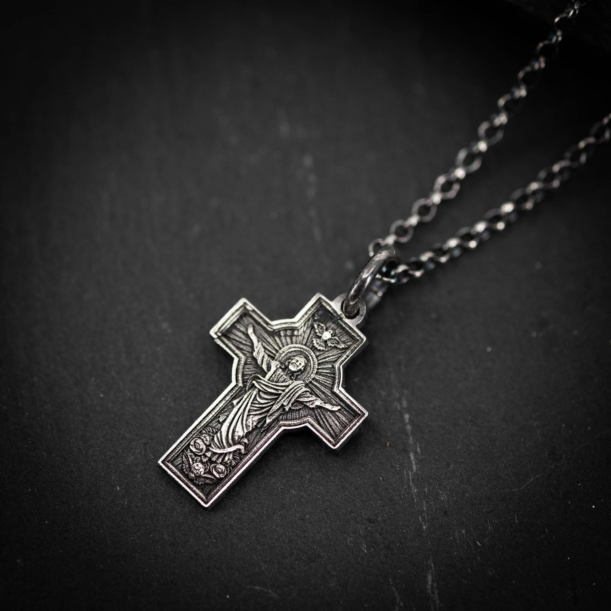 A silver cross pendant with intricate designs and a crown on top, hanging from a chain against a dark background.